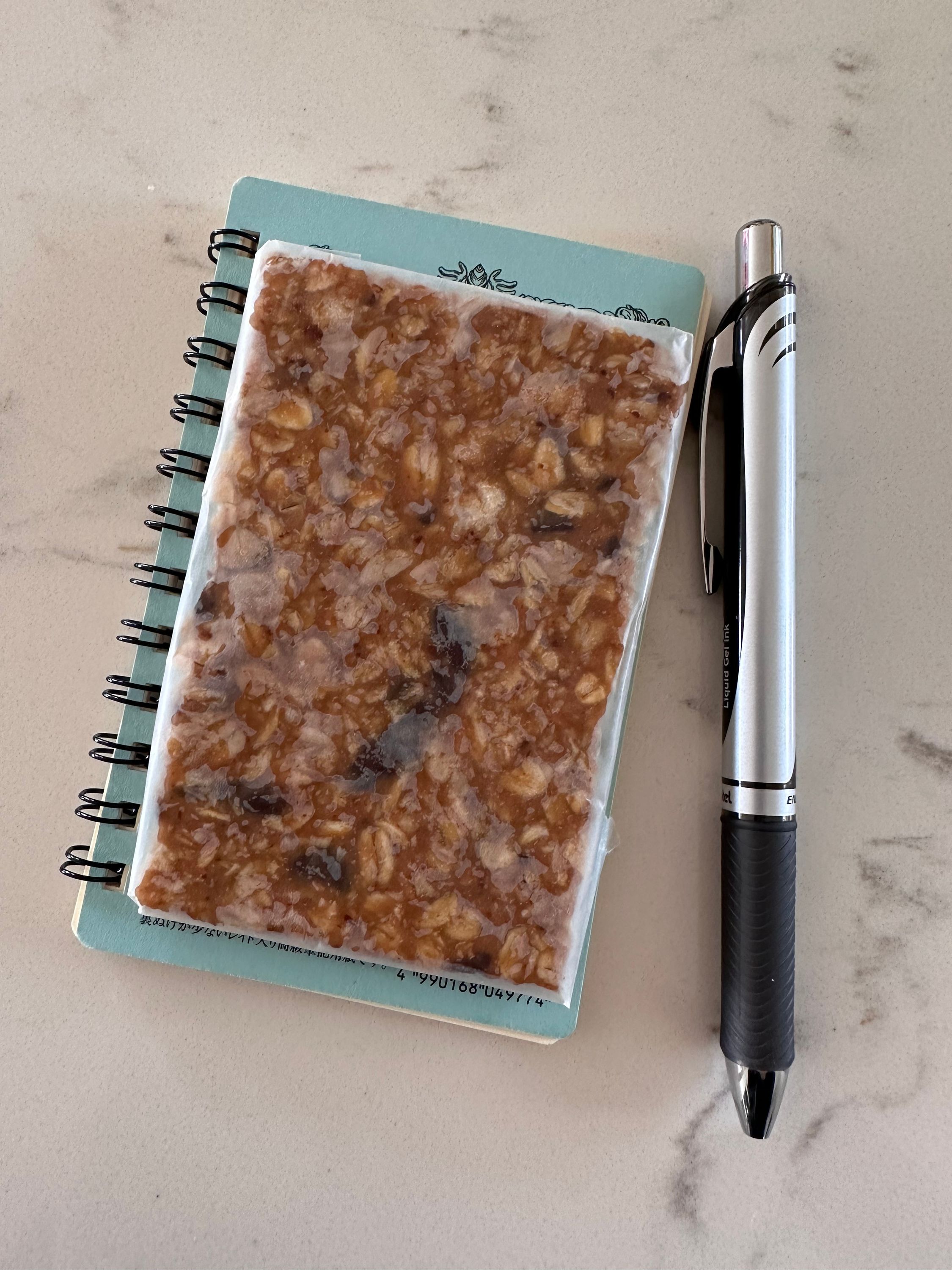 An index card sized granola bar sitting on top of a small pocket notebook next to a Pentel EnerGel pen