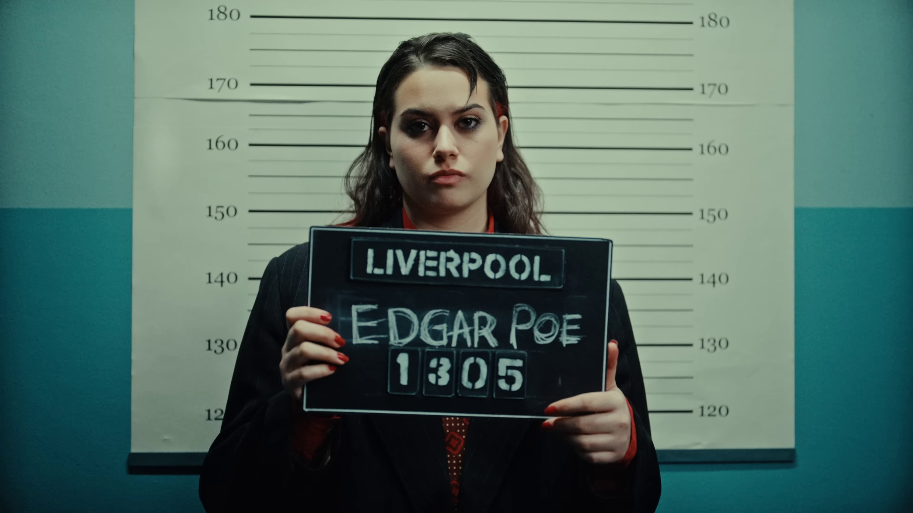 image of Teya holding up a placard that says “Edgar Poe,” as if posing for a police lineup