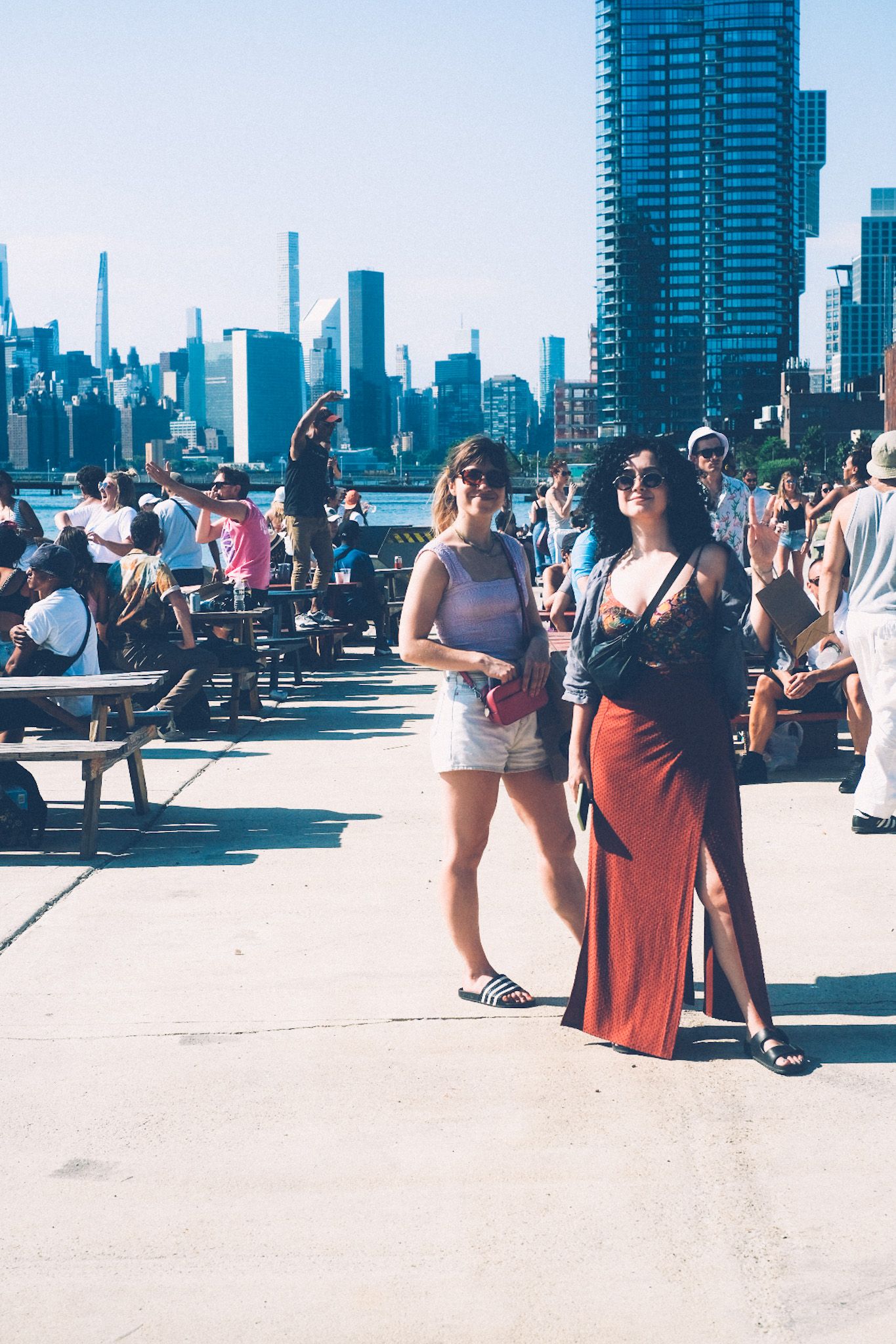 Two women stand on a sunny pier in front of a city skyline, people dancing and sitting on picnic tables in the background.