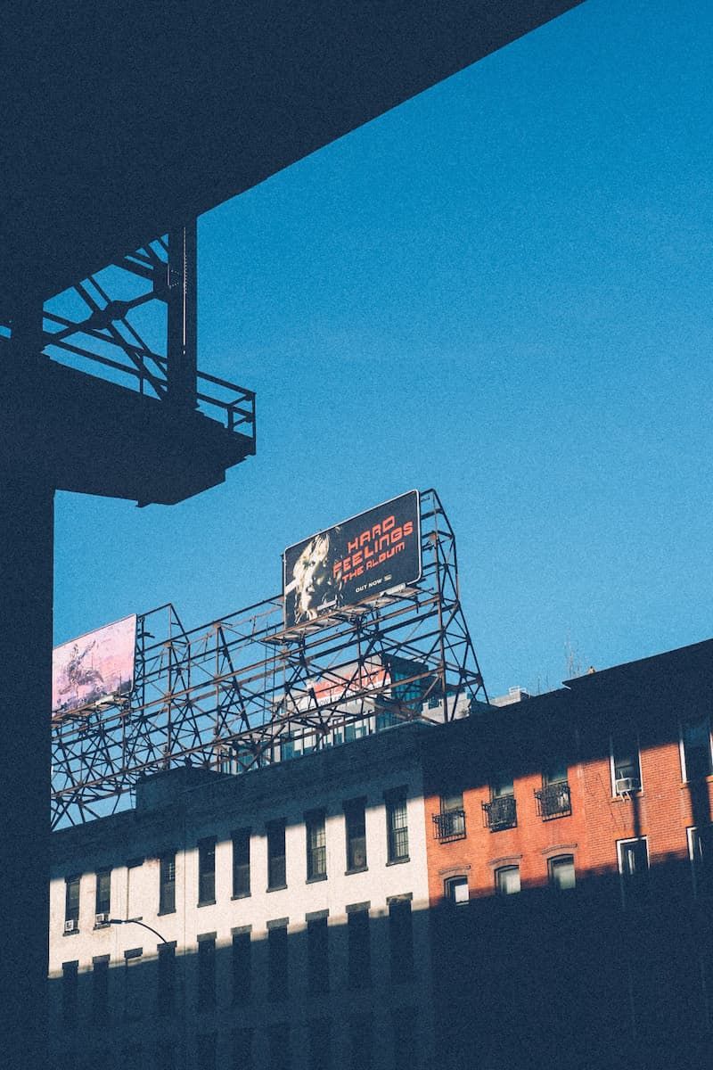 A white brick and red brick building stand in the sunlight under a blue sky, lines of shadow from the bridge crossing the frame. A billboard with the words “Hard Feelings” stands above all.