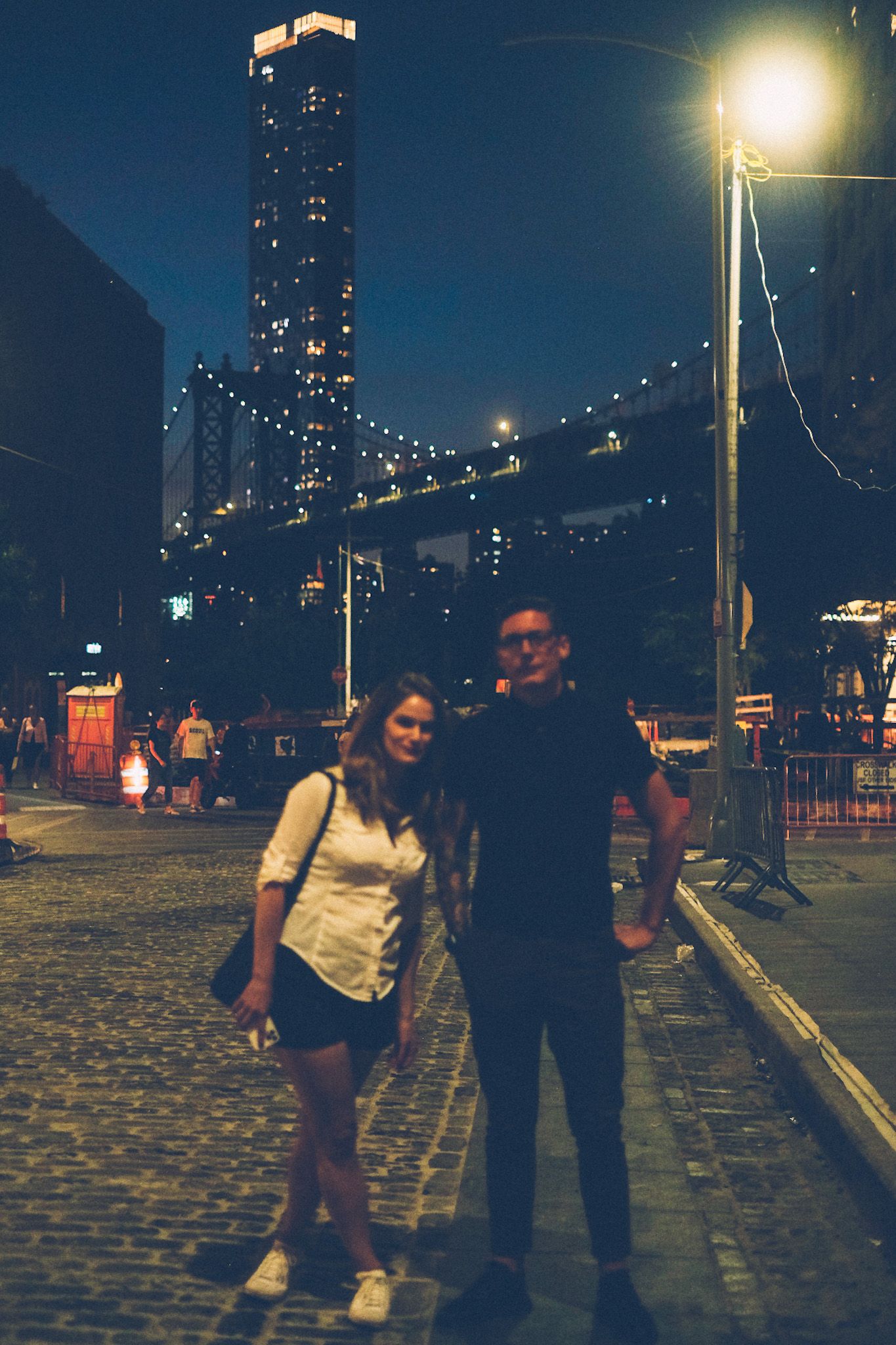 A man and a woman stand out of focus, in front of the Manhattan bridge. It is nighta fun yellow streetlight floods the construction site behind them. They are morose.