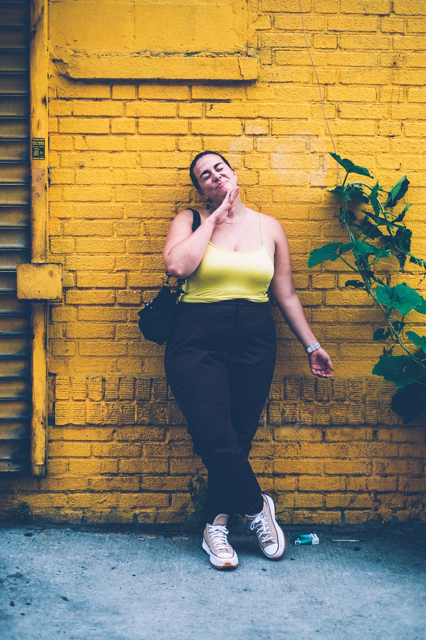 A woman in a yellow tank top and brown pants stands against a vibrant yellow brick wall, a green plant snaking up beside her.