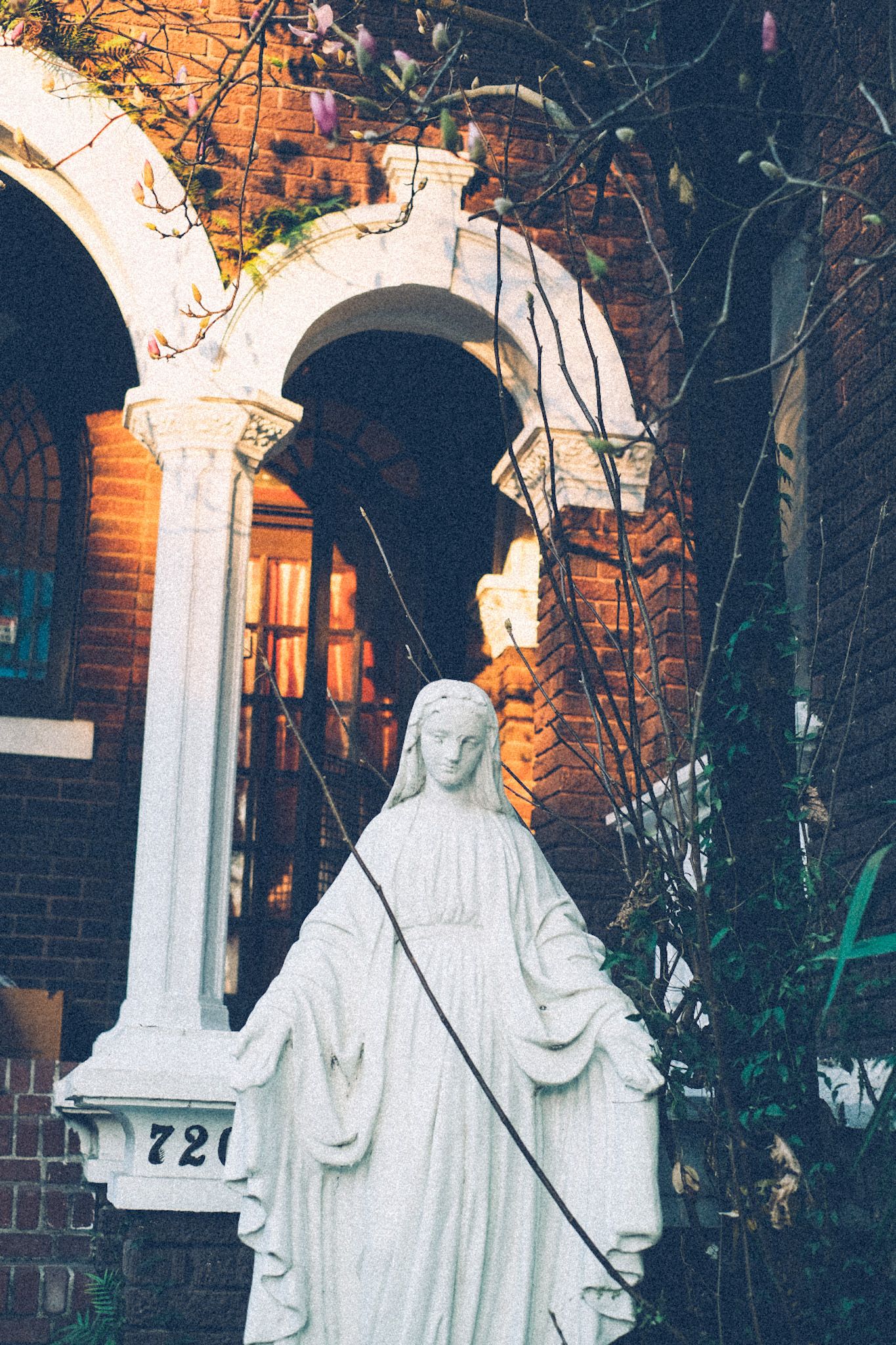 A statue of the Virgin Mary stands in front of an old, beautiful brick house, patches of afternoon light seen in the background.