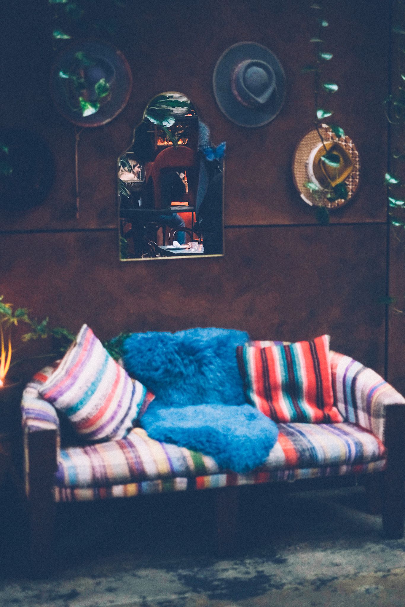 A colorful couch sits in front of a dark brown wall with an antique mirror, wide brim hats and plants all hung on it.
