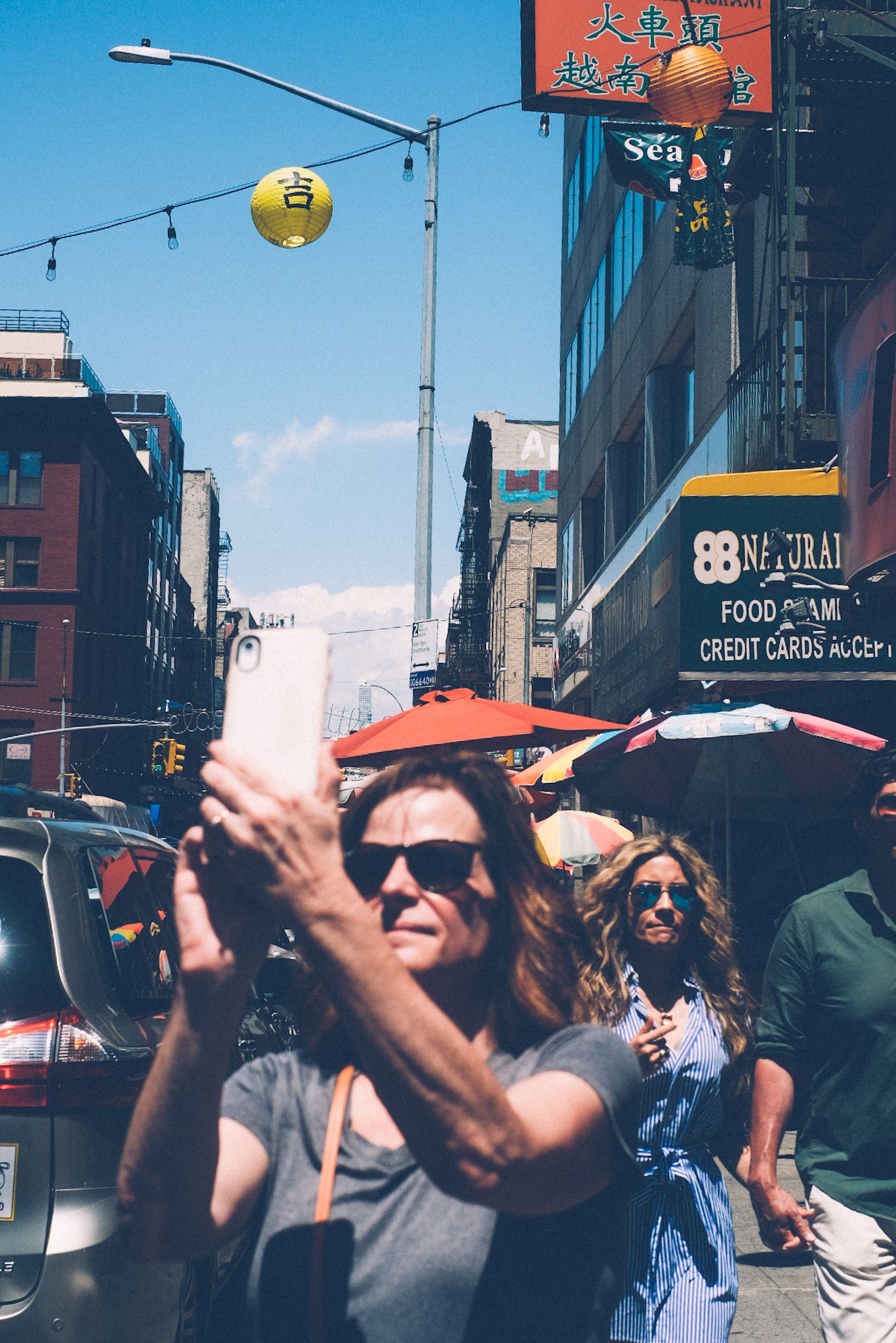 A woman in sunglasses, slightly out of focus, takes a photo with her phone in Chinatown. Other people pass behind her. The sky is so blue.