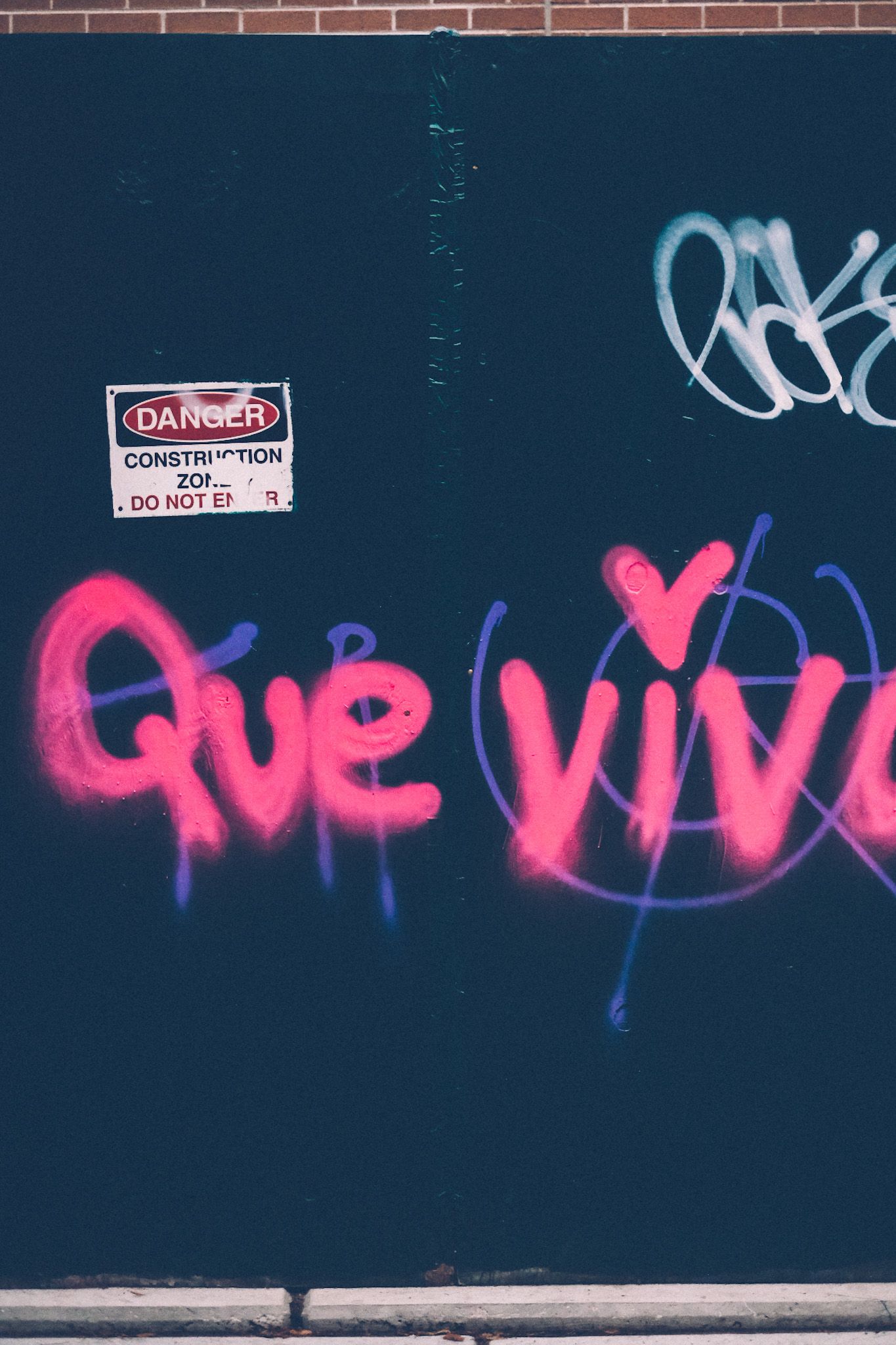 “Que vive” is spray painted in neon pink against a dark temporary construction wall.