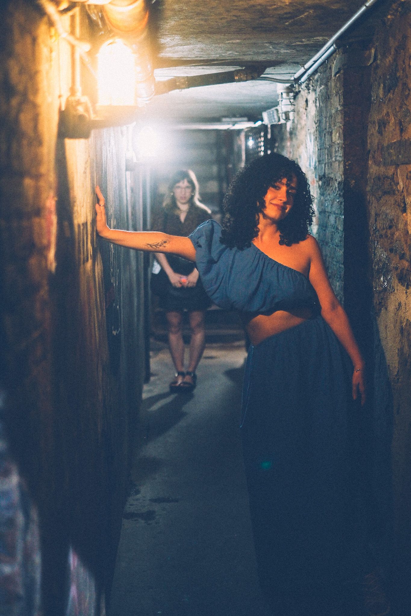 Two women stand in a dark alleyway lit by two lights, one warm and one cooler. In the front, the woman wears a blue dress, in focus, her arm across the alleyway and hand against the brick wall. In the back, the second woman stands out of focus, hands together near her lap.