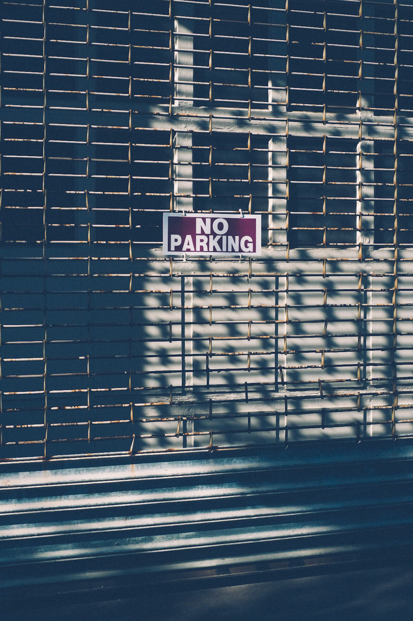 A burgundy sign saying “no parking” is placed in the center of a white grated shutter of a storefront. The afternoon shadow is lovely.