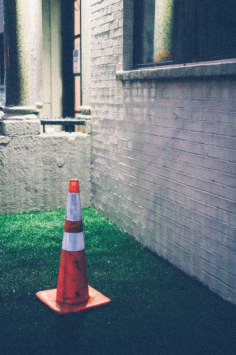 An orange construction cone sits in the corner of a patch of grass, under a streetlight against white brick.