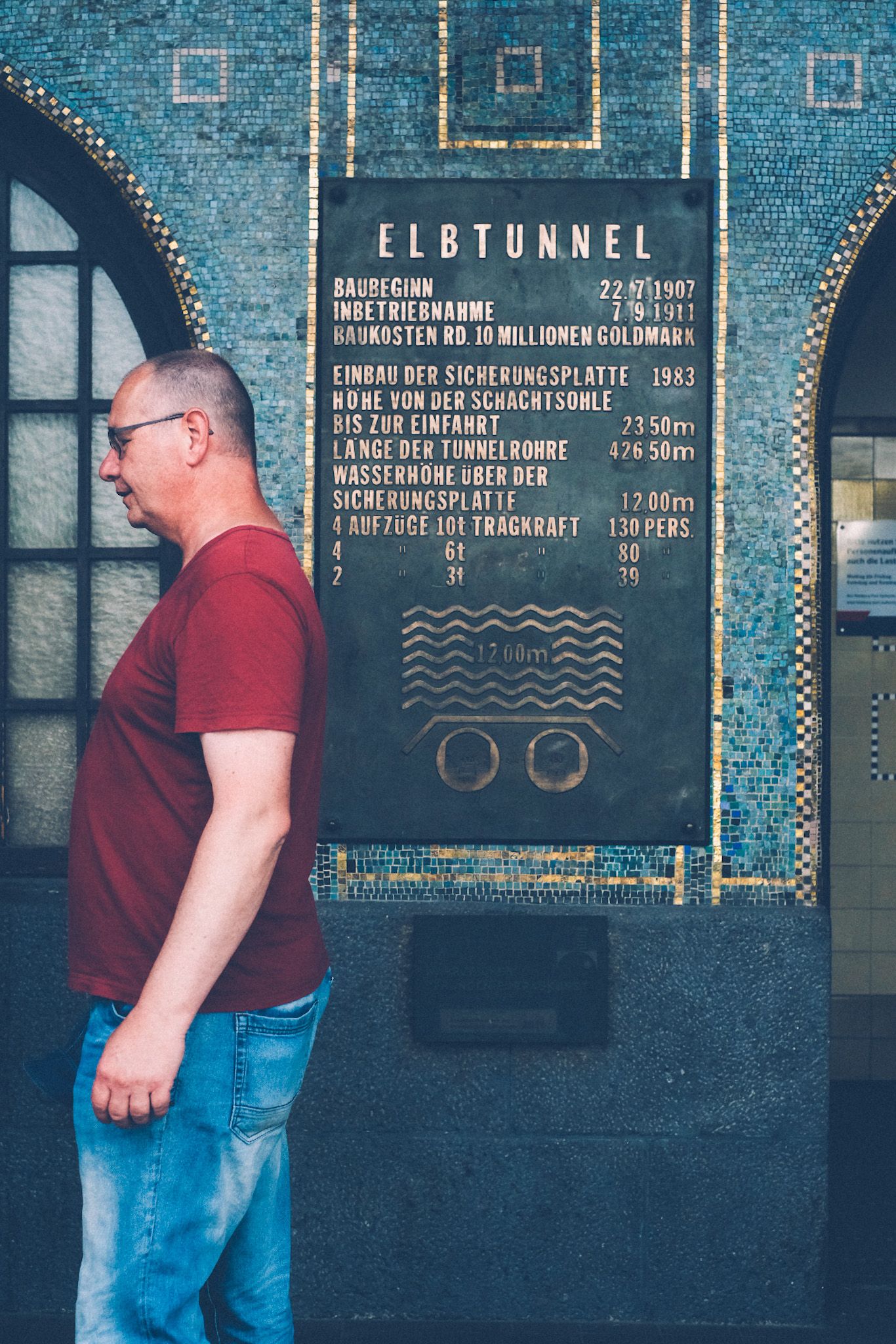 A man in a red shirt stands with his back to a sign saying “Elbtunnel” tiled onto a wall in Hamburg, Germany.
