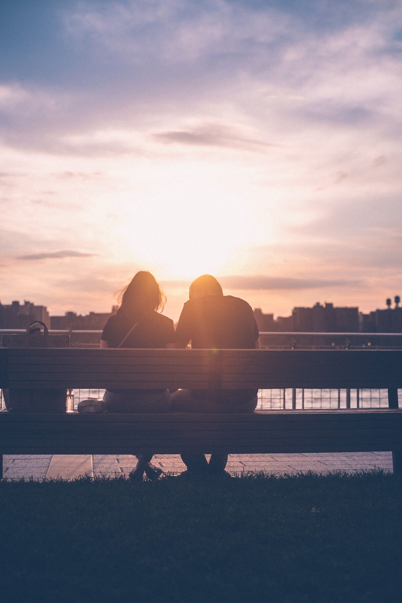 The silhouette of a couple leaning toward one another in front of a warm sunset, sitting on a bench by water.