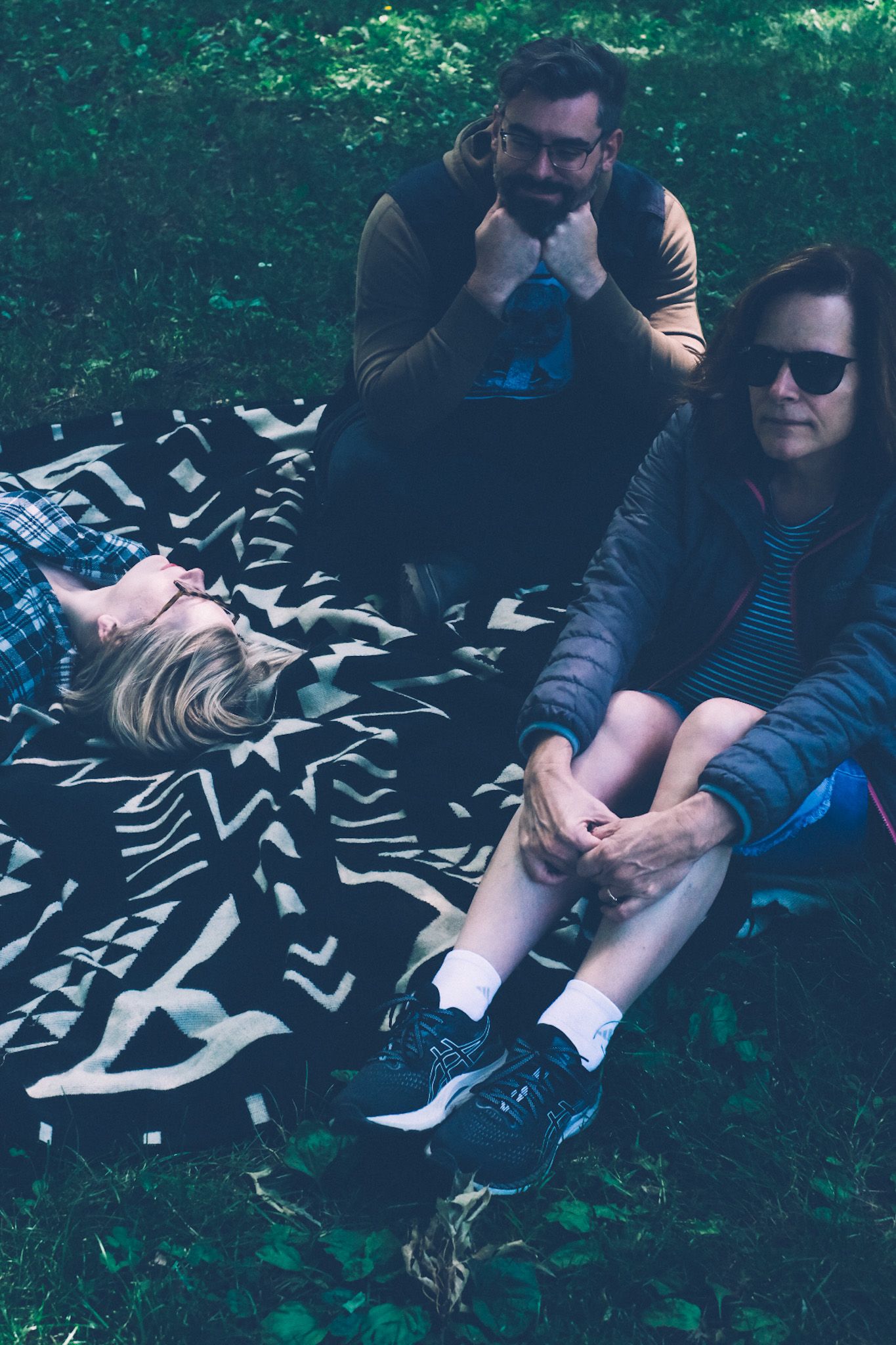 Three people sit on a black and white patterned blanket in the grass.