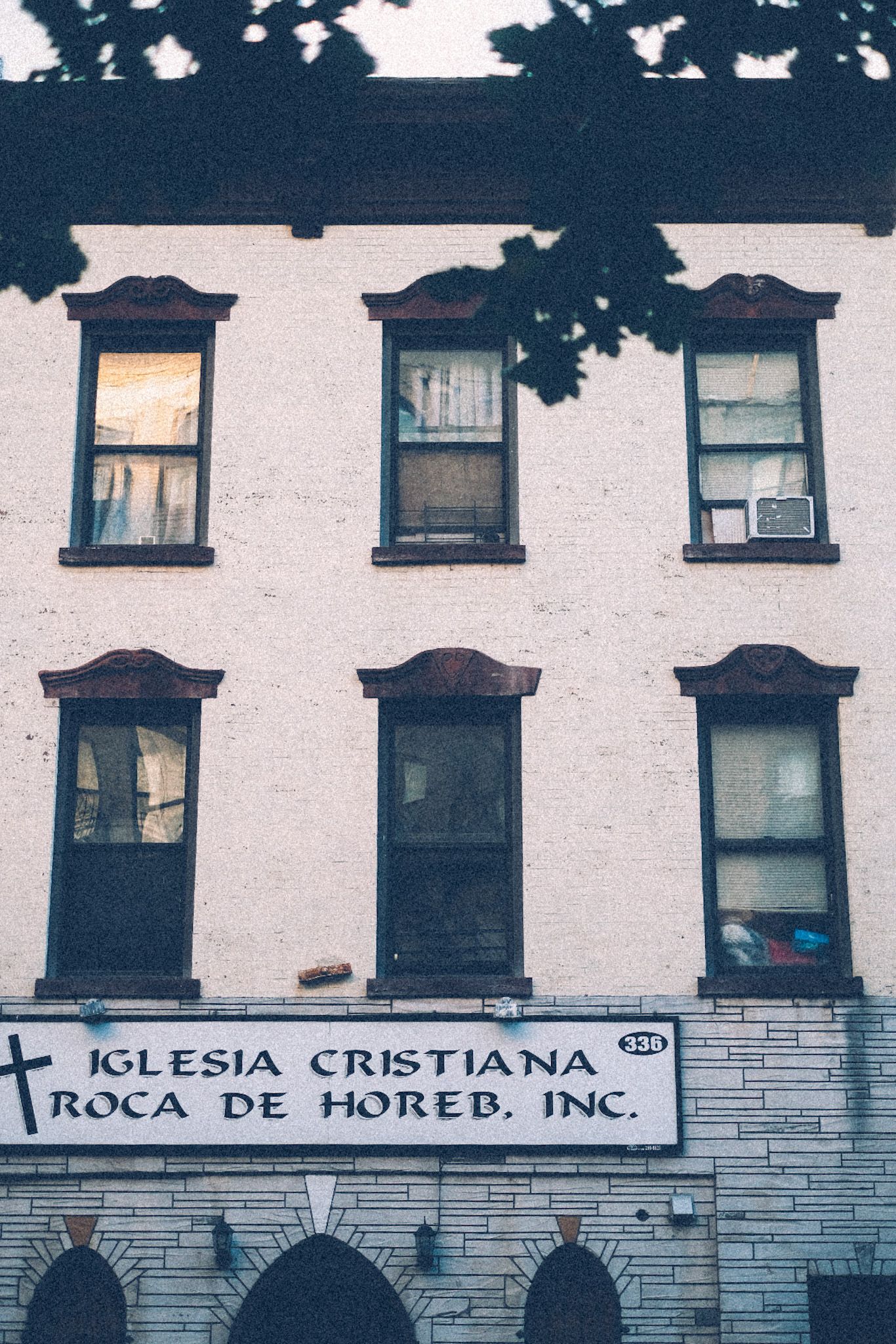 A white three-story building with dark brown trim roses up from the bottom of the photo, with a hand-painted sign saying “Iglesia Cristiana”.