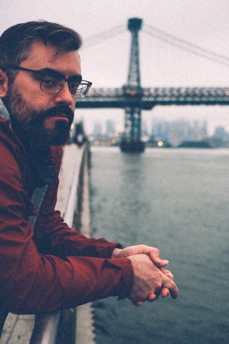 A dark haired man standing stoically on the edge of the water on an overcast day, a bridge in the background