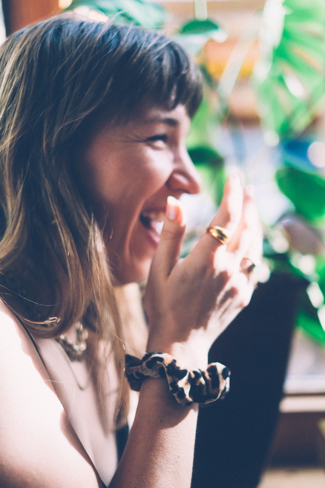 A close-up side profile of a woman laughing with her hand up, wrist wrapped with a leopard print scrunchie. Natural light floods in from the window in the background, plants standing just out of focus.
