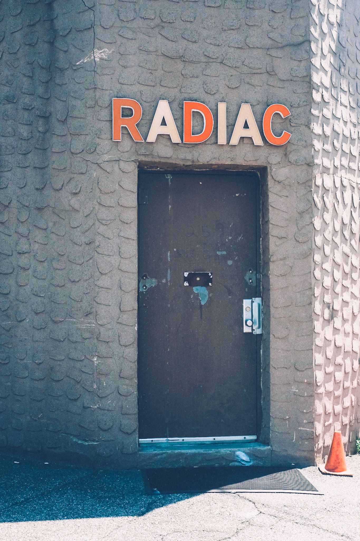 A doorway on the corner of a grey wall has big white and orange letters saying RADIAC. The wall is textured almost like scales, lit in sun but also in shadow. An orange cone sits to the right of the door.