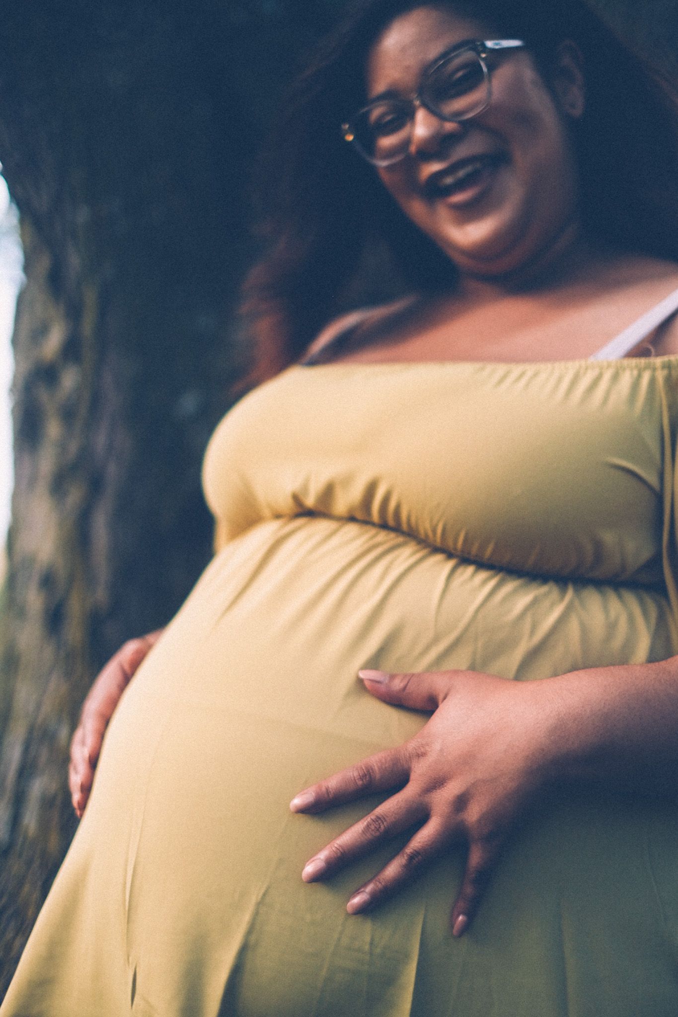 A pregnant woman in a yellow dress smiles and holds her belly.