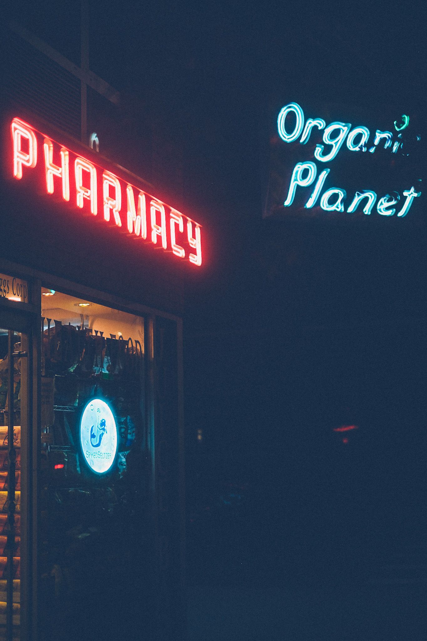 Two neon signs glow from the darkness. One says organic planet, in blue, while the other days Pharmacy in red over a doorway in the dark.