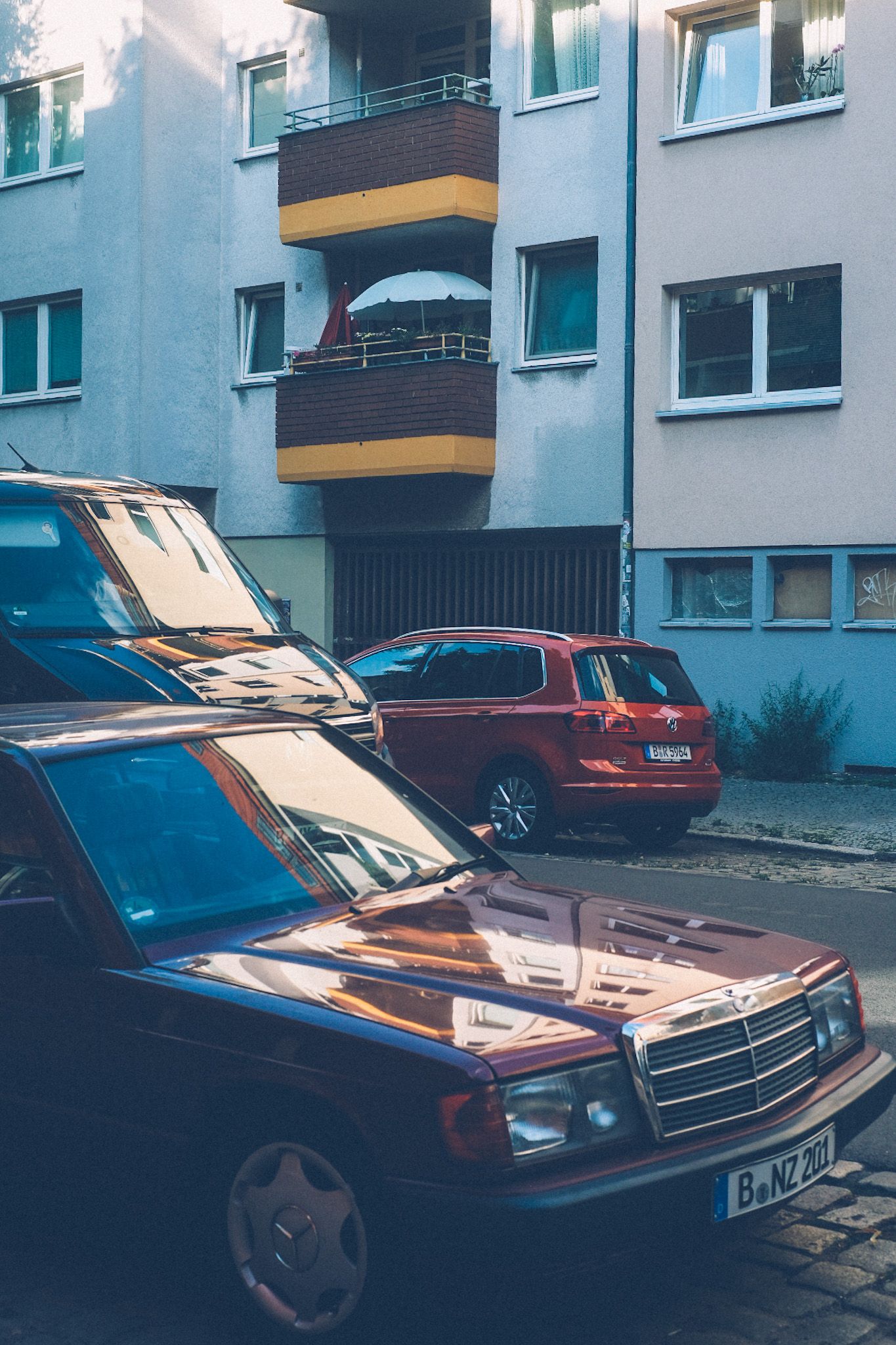 Three cars overlap, parked in a street in Berlin, in front of a white apartment building with balconies that are brown and yellow.