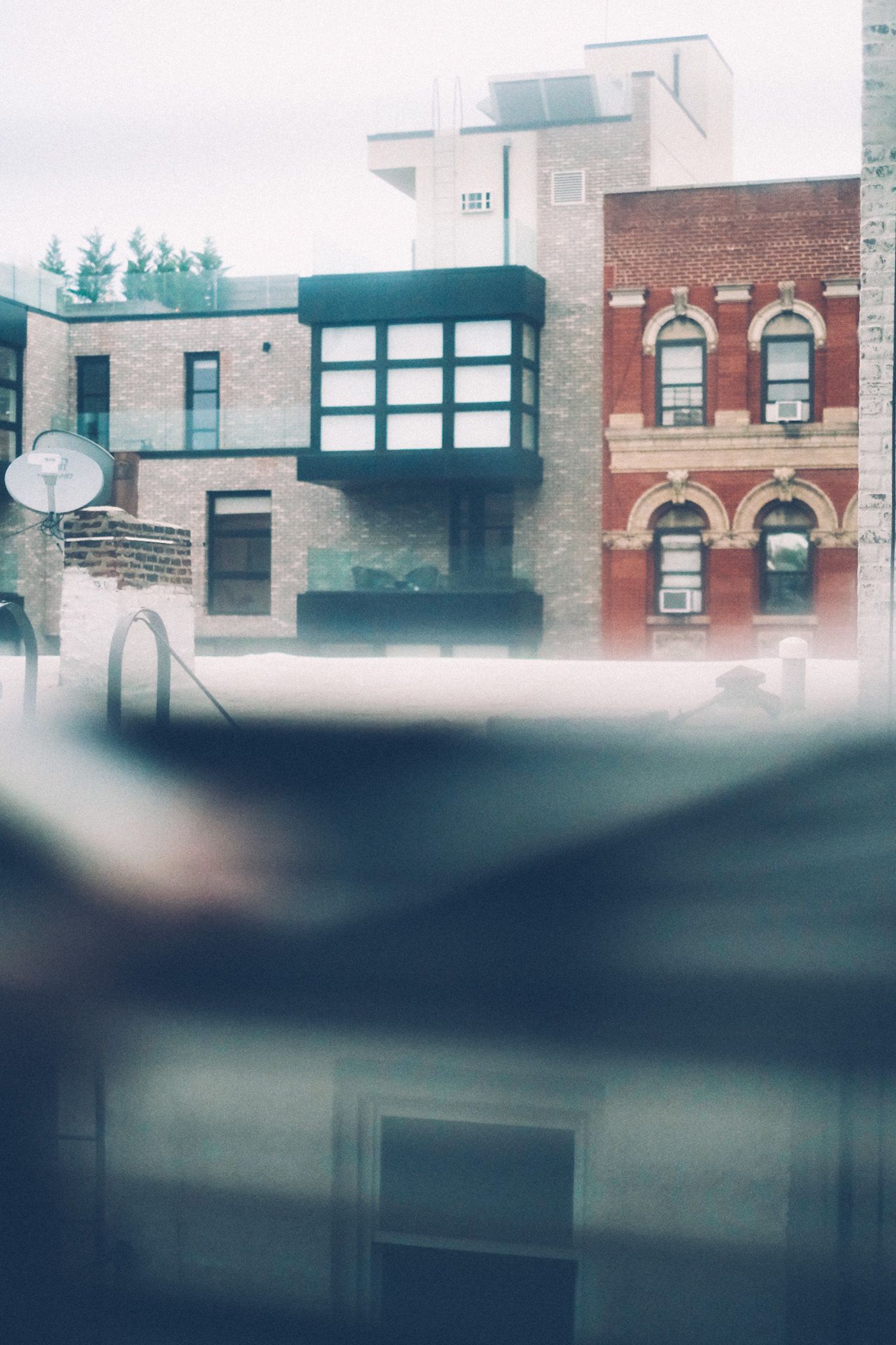 Red and white brick buildings are seen through a window, blind bent downward, on an overcast day.