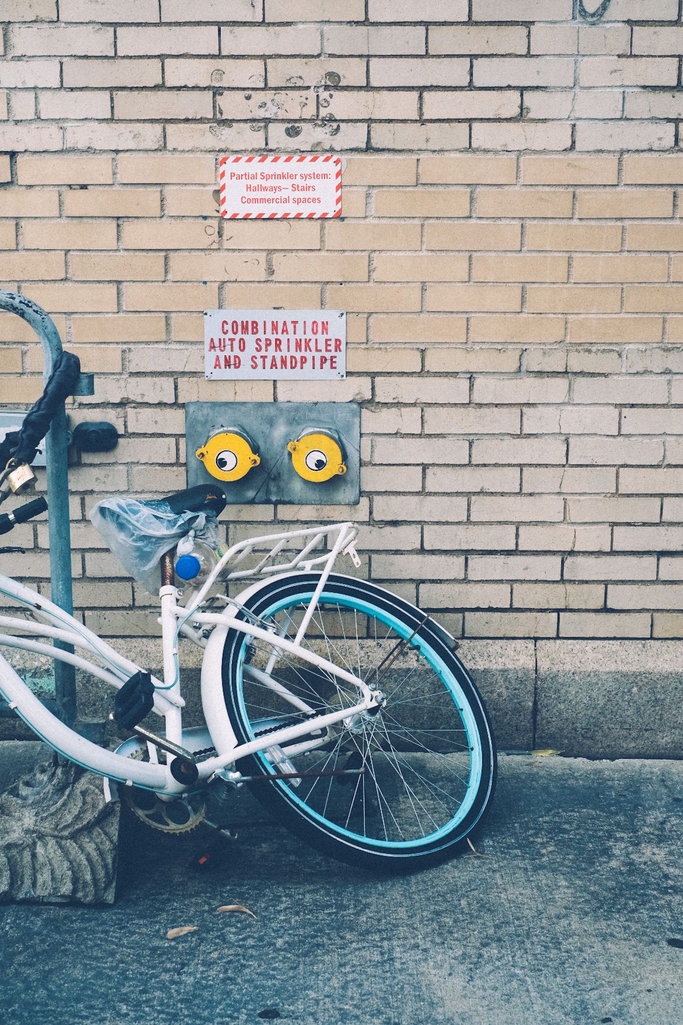 A white bike leans against an off-white brick wall, in front of a sprinkler hookup with googly eyes on it.