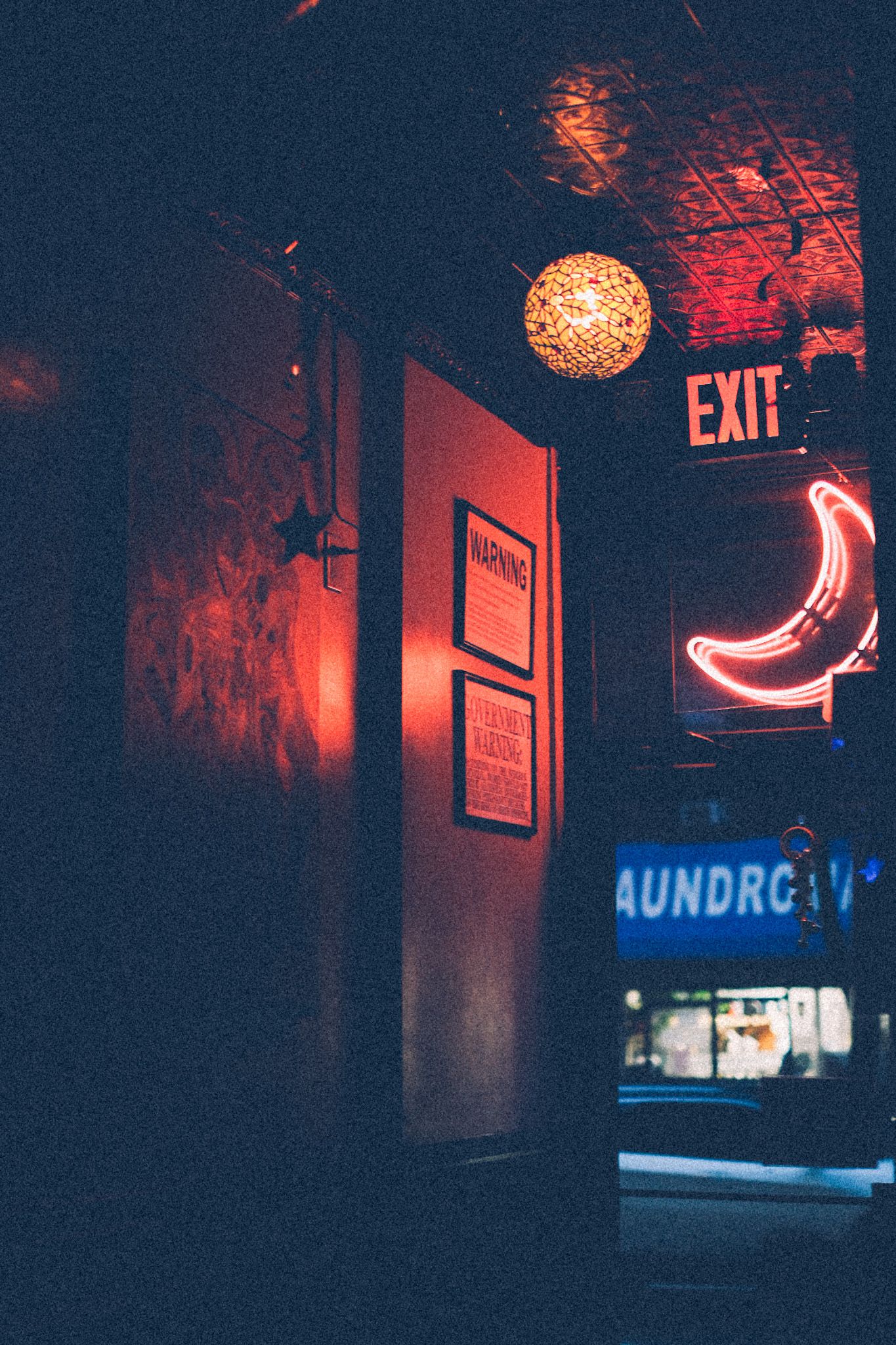 The doorway of a bar glows at night, lit by a red neon sign in the shape of a moon and an exit sign