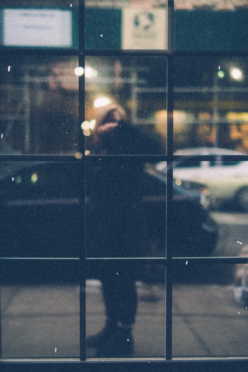 A paned window reflection of a woman in a dark coat on a city street at dusk, scaffolding and cars behind her.