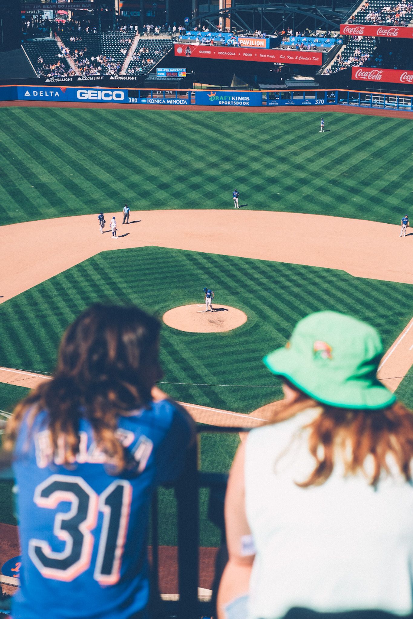 The backs of two women at a baseball game frame the pitcher’s mound of a major league baseball field on a sunny day.