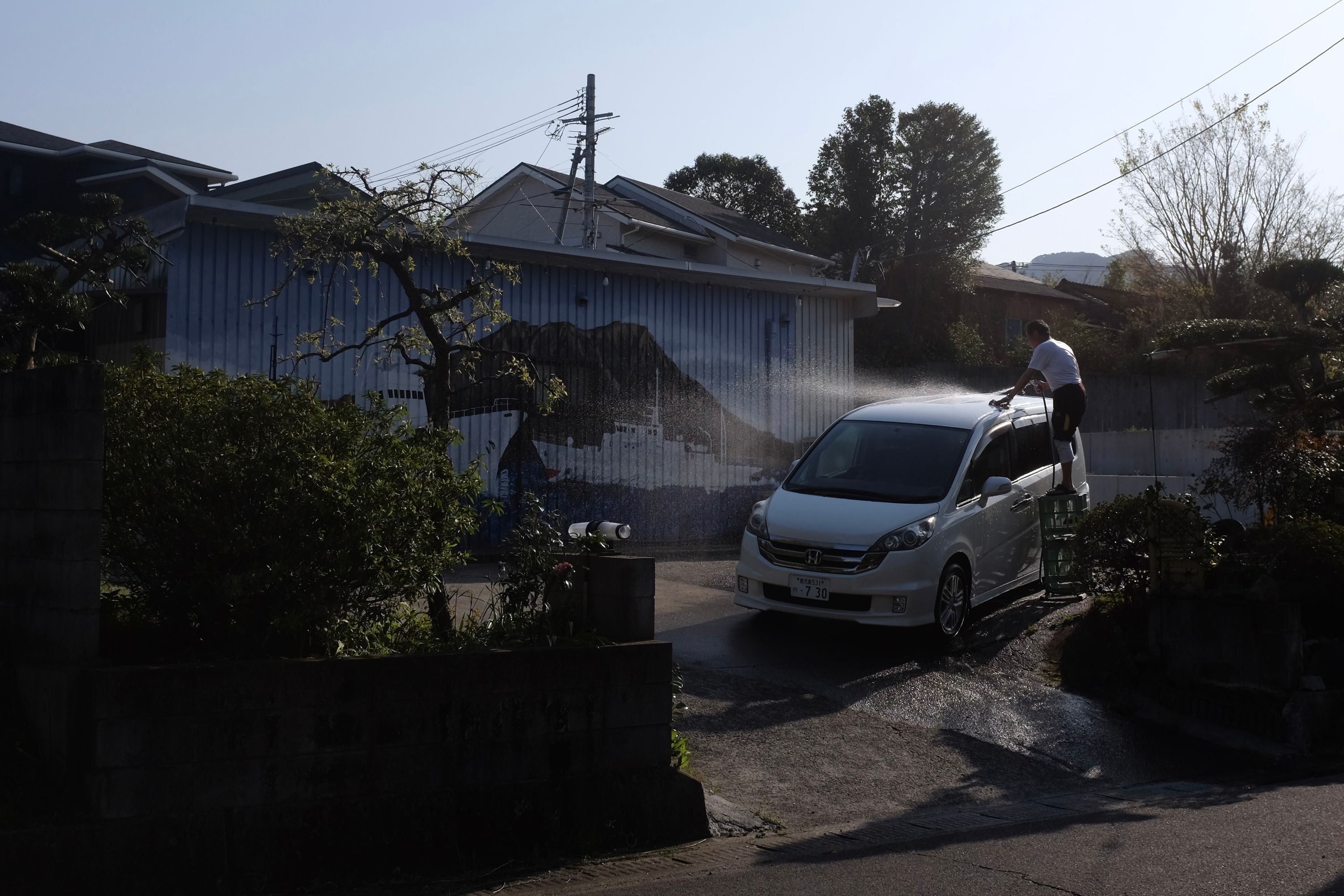 A man washes his minivan in front of a garage with the volcano Sakurajima painted on its door.