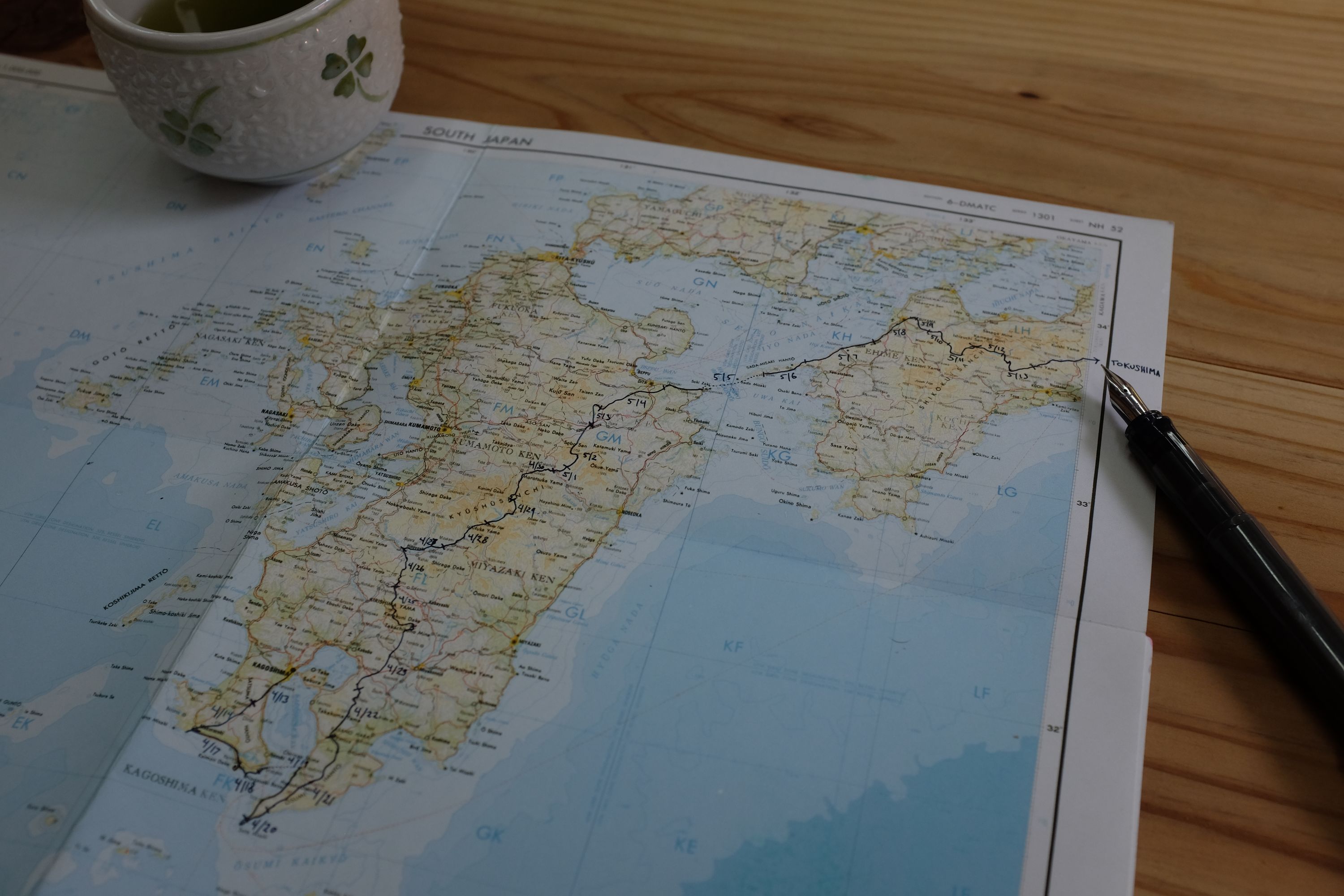 A paper military map of South Japan held down by a teacup, with the route of These Walking Dreams highlighted with a fountain pen; the tip of the pen is visible in the corner.