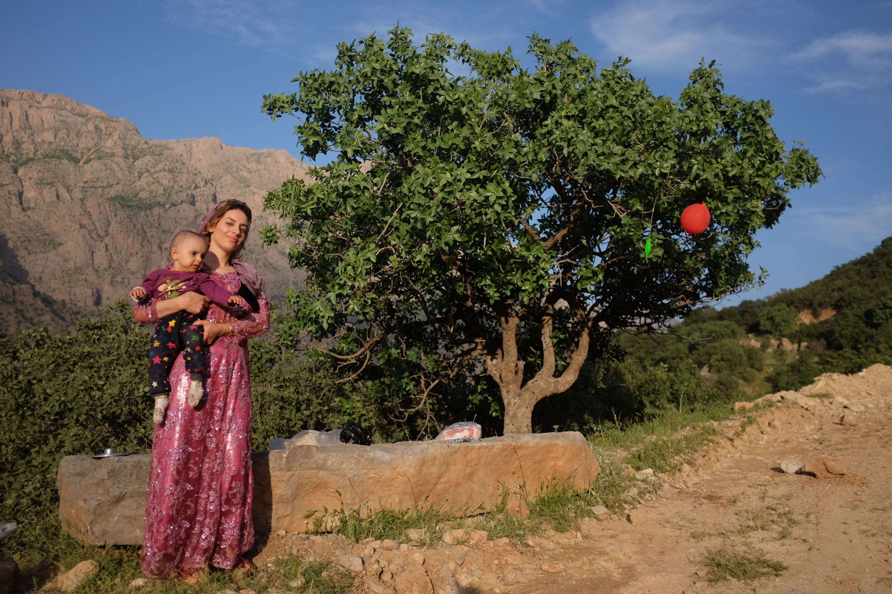 A young woman in an embroidered pink dress holds a baby in front of a large tree with a red balloon in its branches.