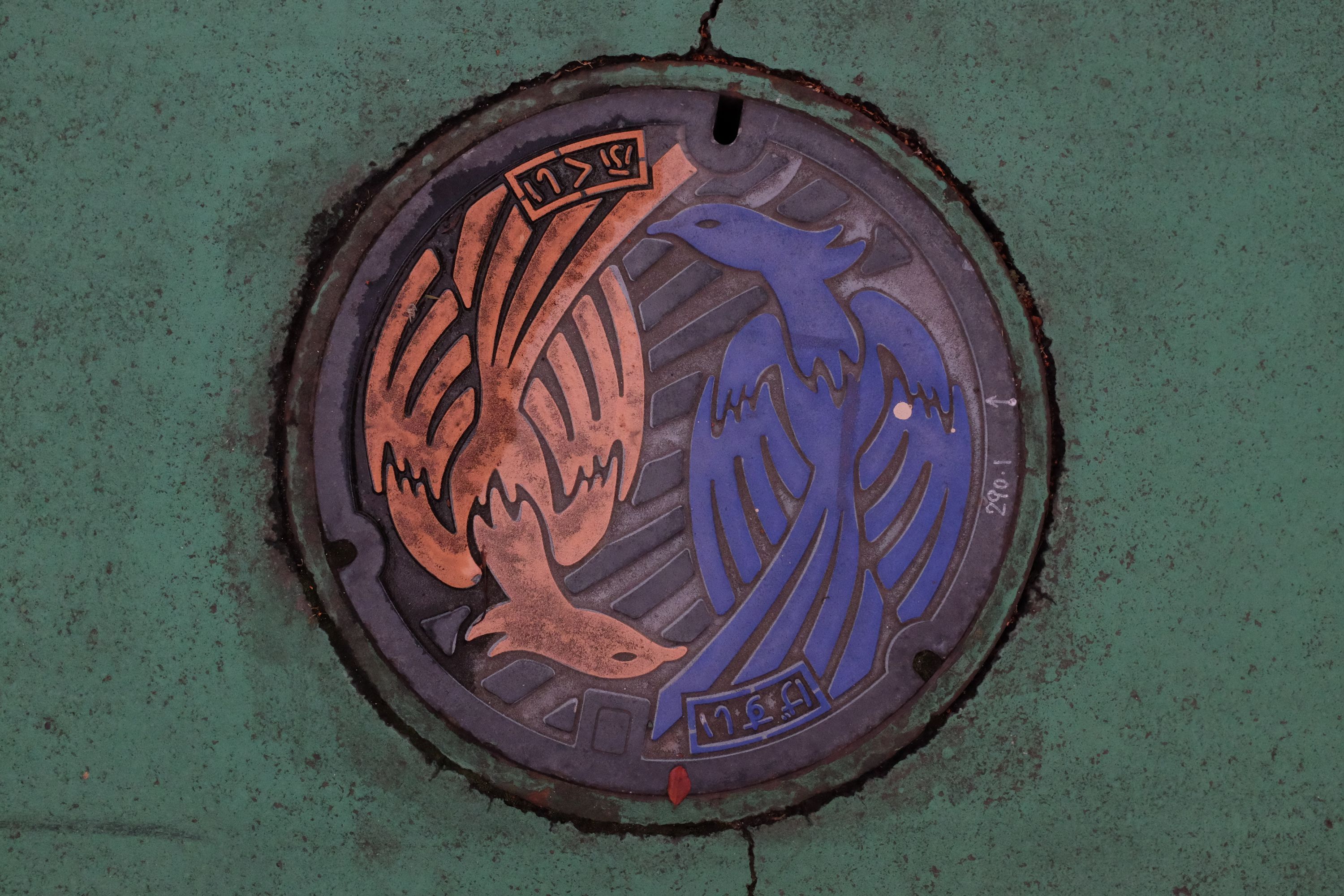 A manhole cover with two dragon-like birds, one yellow, one blue.