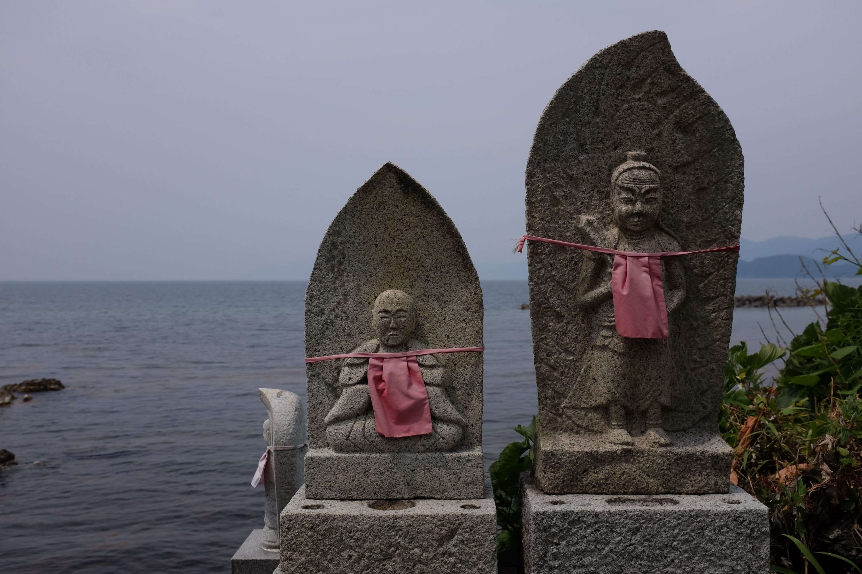 Three small Buddhist statues, wearing red aprons, stand on the shore of the Sea of Japan.
