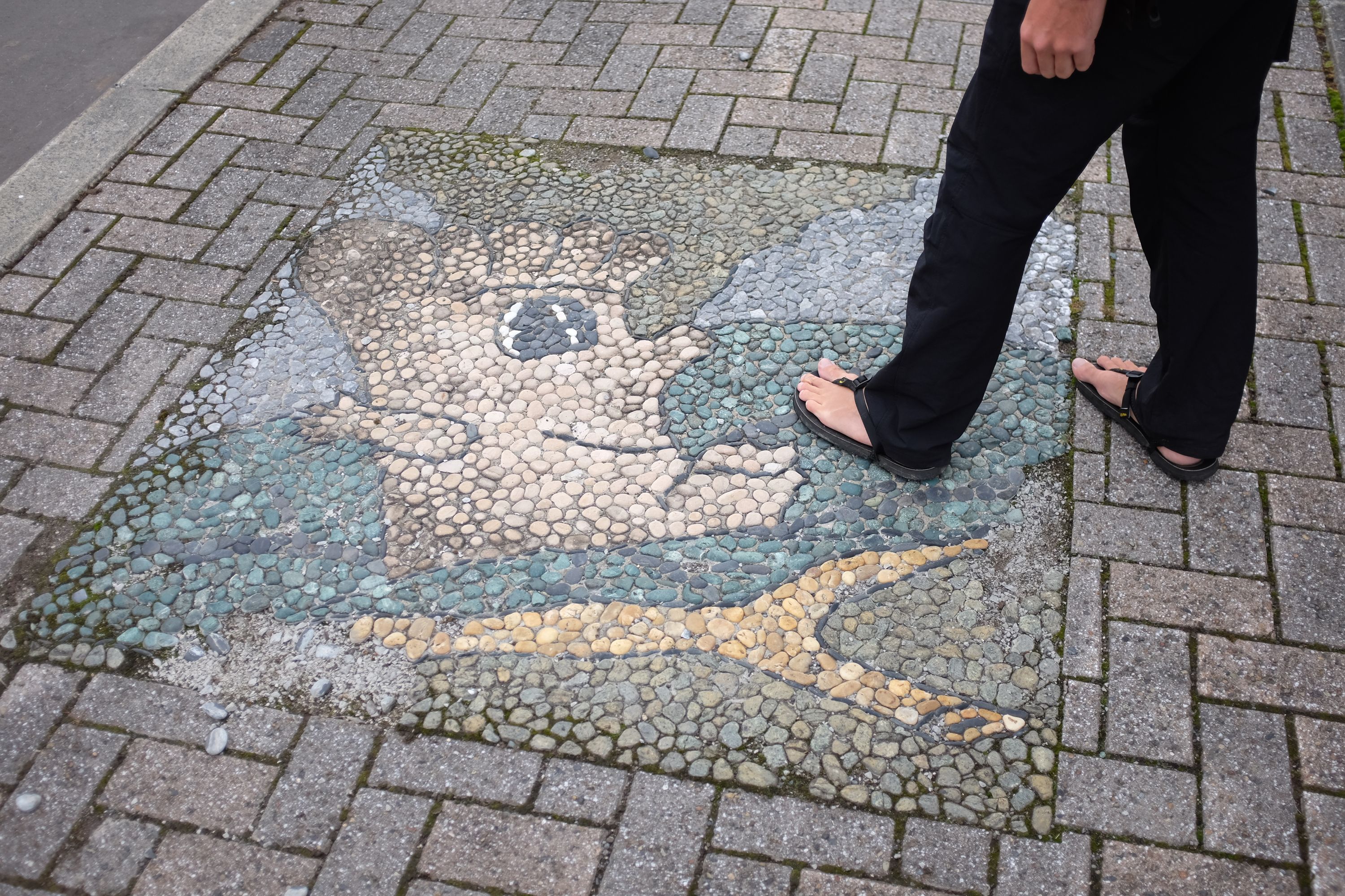 Gabor stands by a mosaic of the foot in sandals, visible from the waist down.