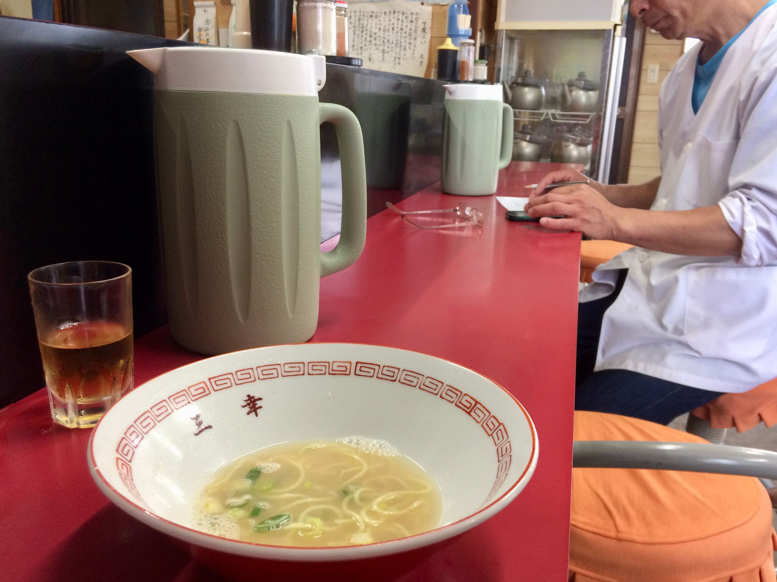 A bowl of half-eaten ramen on the counter of a restaurant, with a man writing a note on a pad behind it.