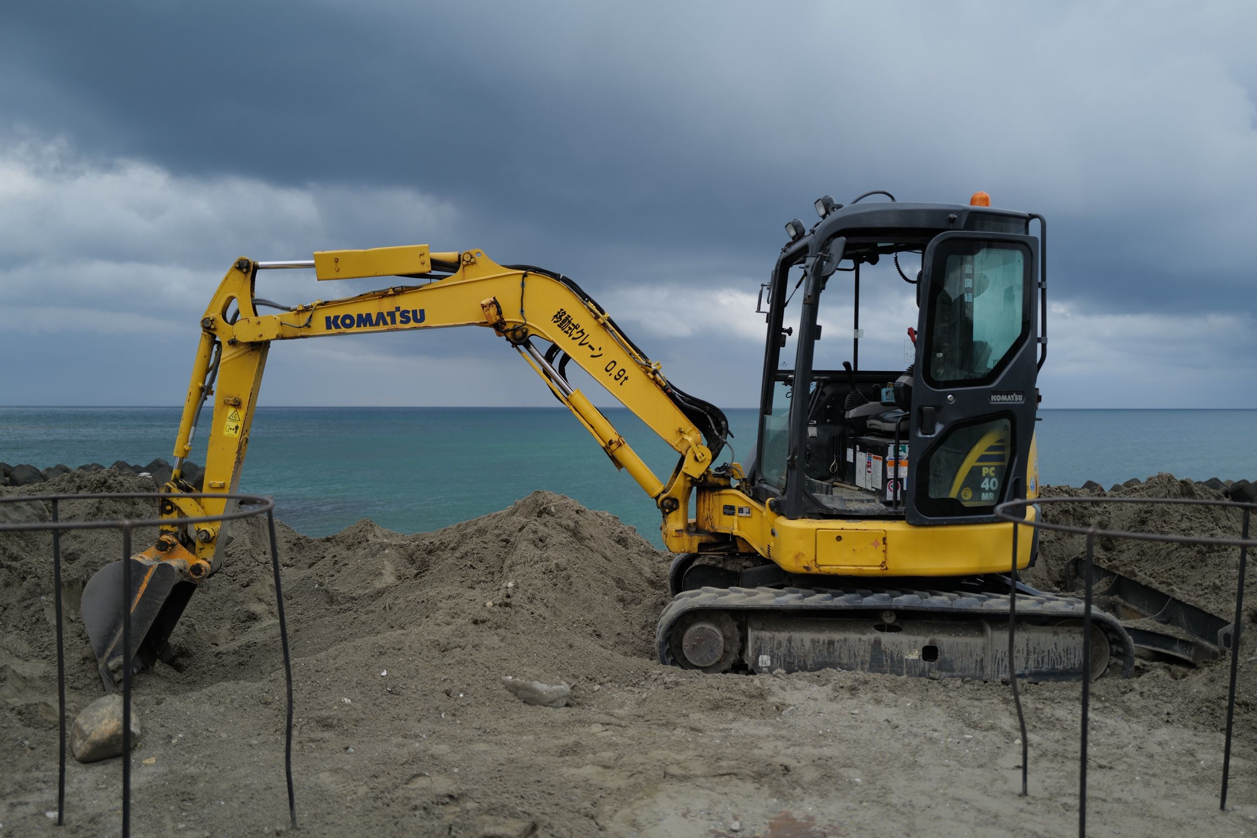 A small yellow excavator stands on a beach under dramatic clouds