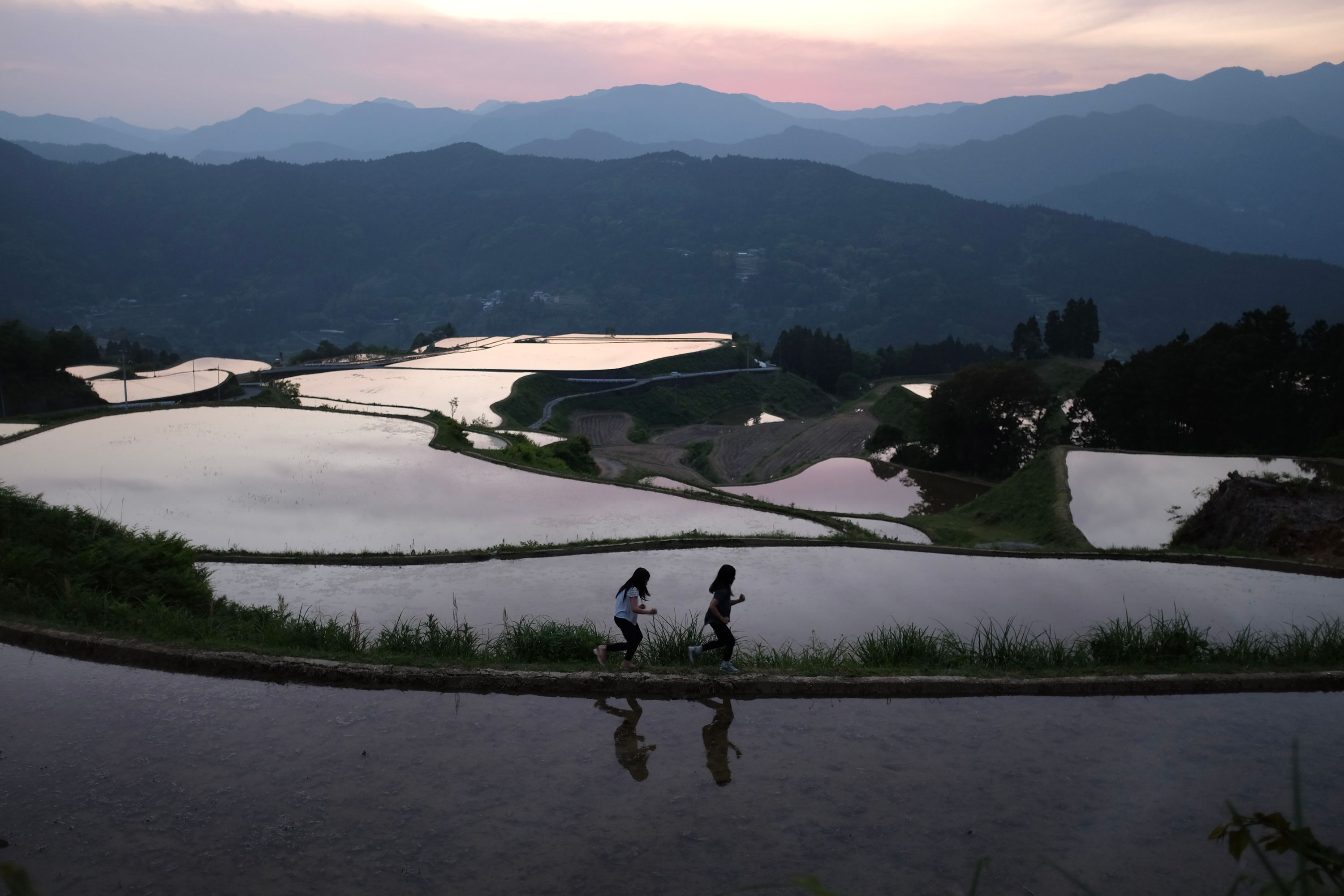 Two young girls run along the dyke between two flooded ricefields, with more ricefields, ranges of hills, and the setting Sun visible behind them.