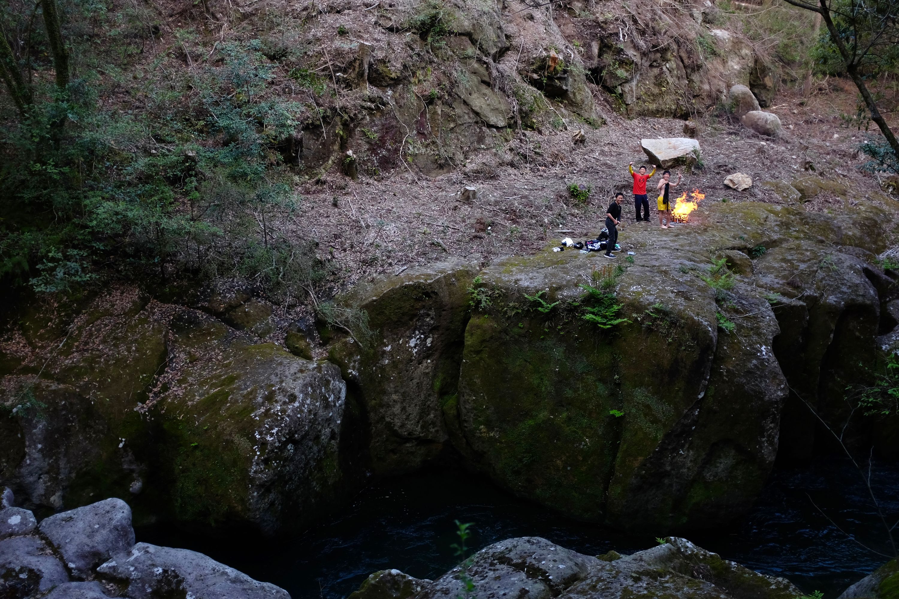 Kids waving by a campfire on the opposite bank of a stream with steep black rock walls.