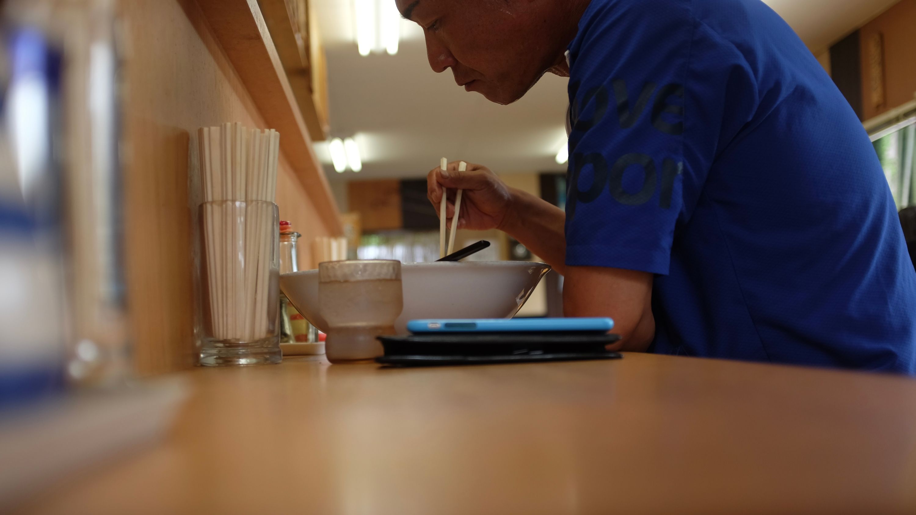 A man in a blue t-shirt sits at a counter and eats a similar bowl of ramen.