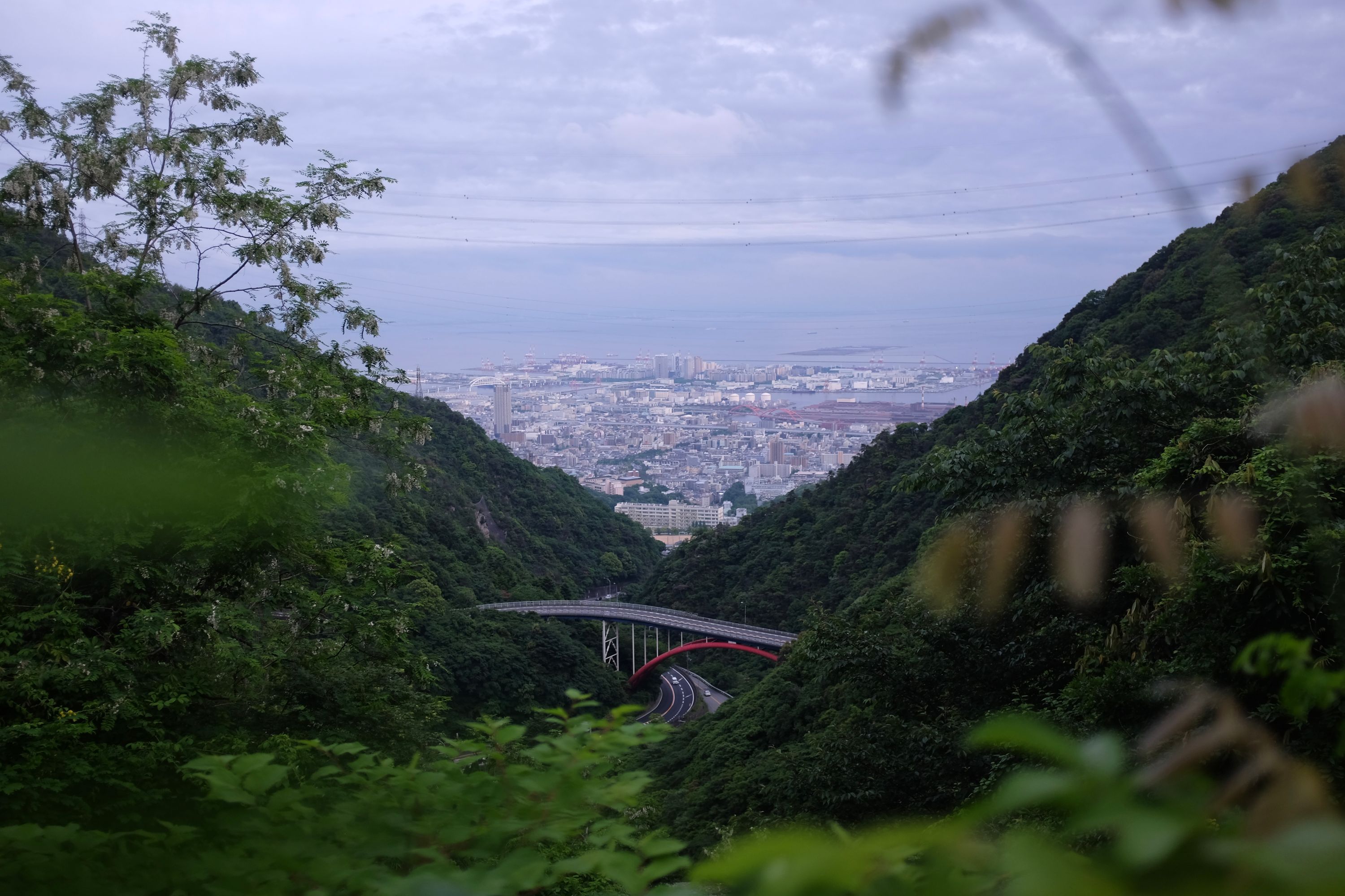 Looking down on a twisting mountain road, then Kōbe, then the sea beyond, under an overcast sky.