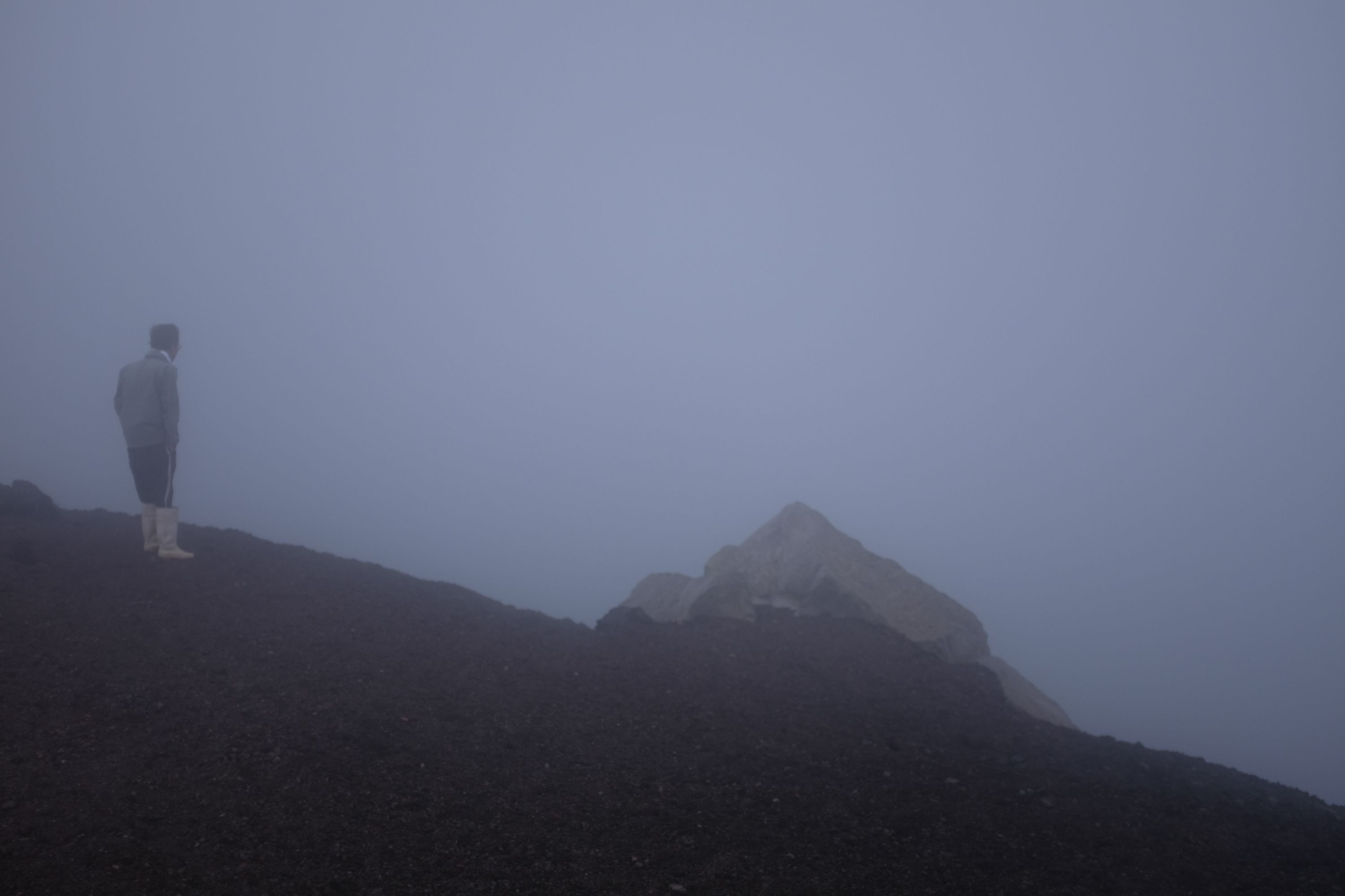 A man in white rubber boots, standing on a scree slope, looks into the fog.