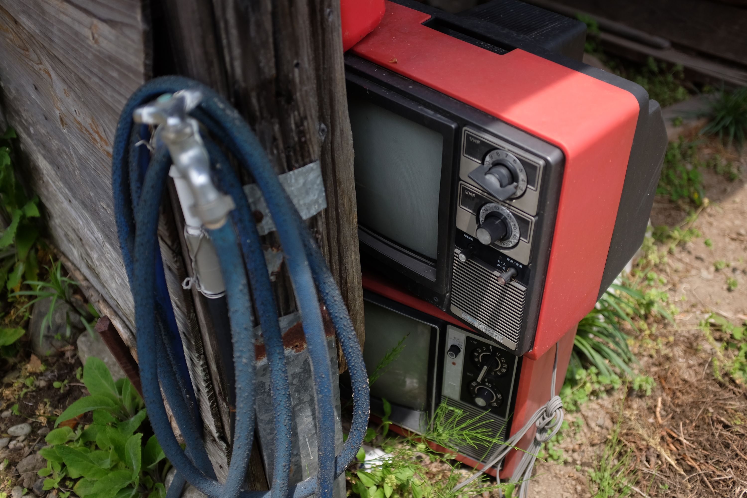 A coiled-up blue hose and two old red televisions in a shed overgrowing with weeds.