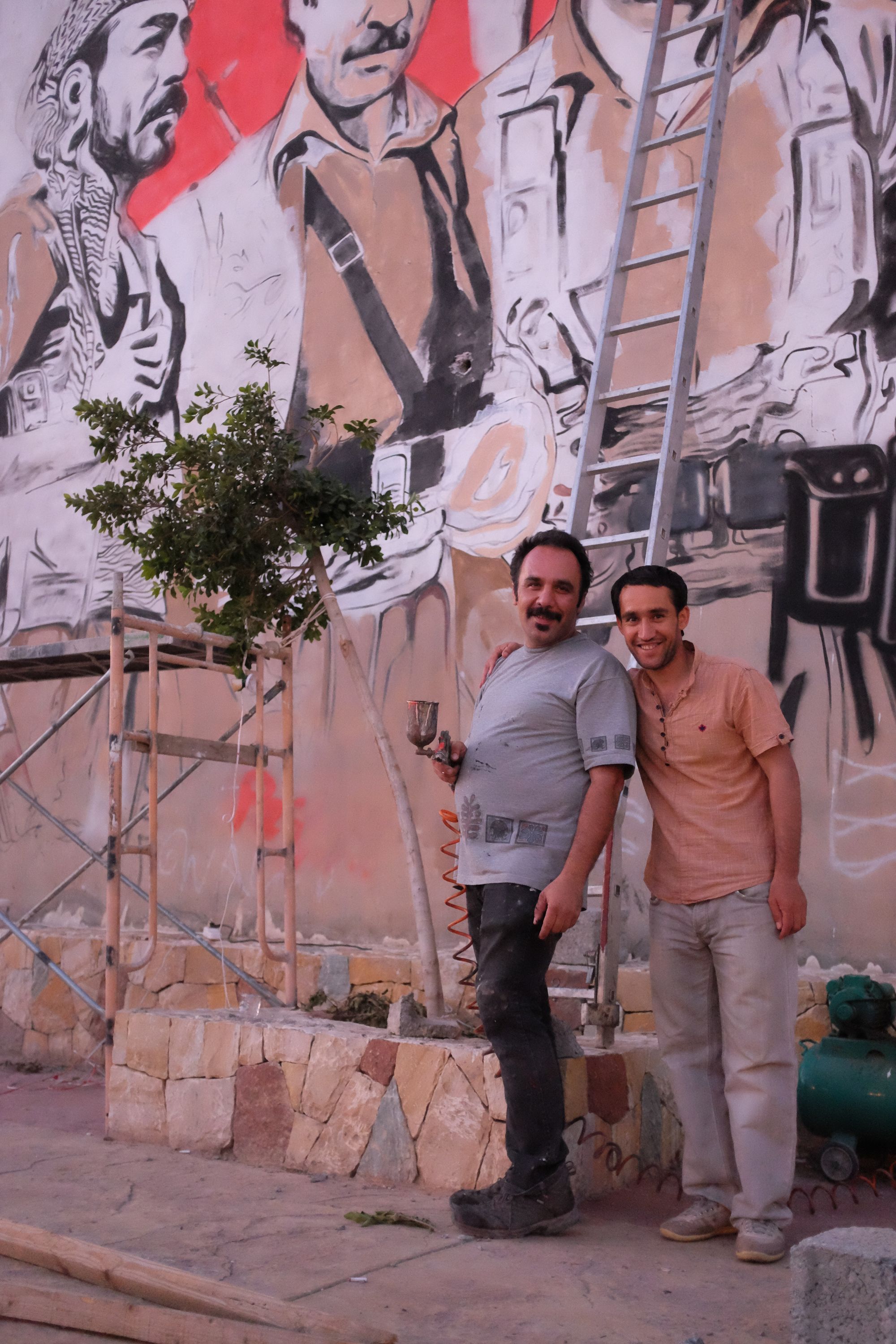 Two men with spray paint guns smile into the camera in front of a large mural which depicts peshmerga, Kurdish warriors.