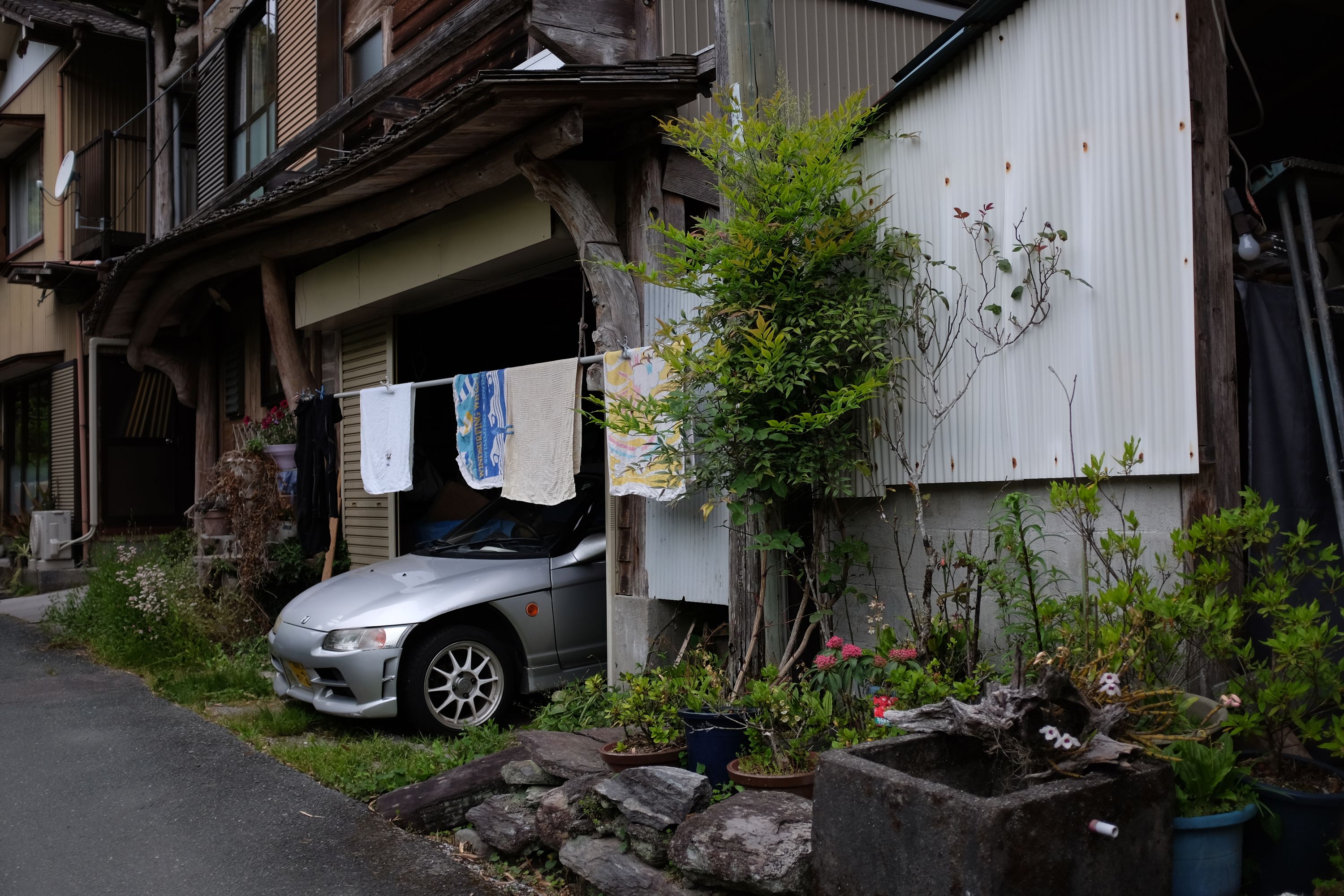 A Honda Beat roadster peeks out of a garage in a village house, the kind with hand-made windowframes, a small rock garden in front, and laundry hung across the garage’s entrance.