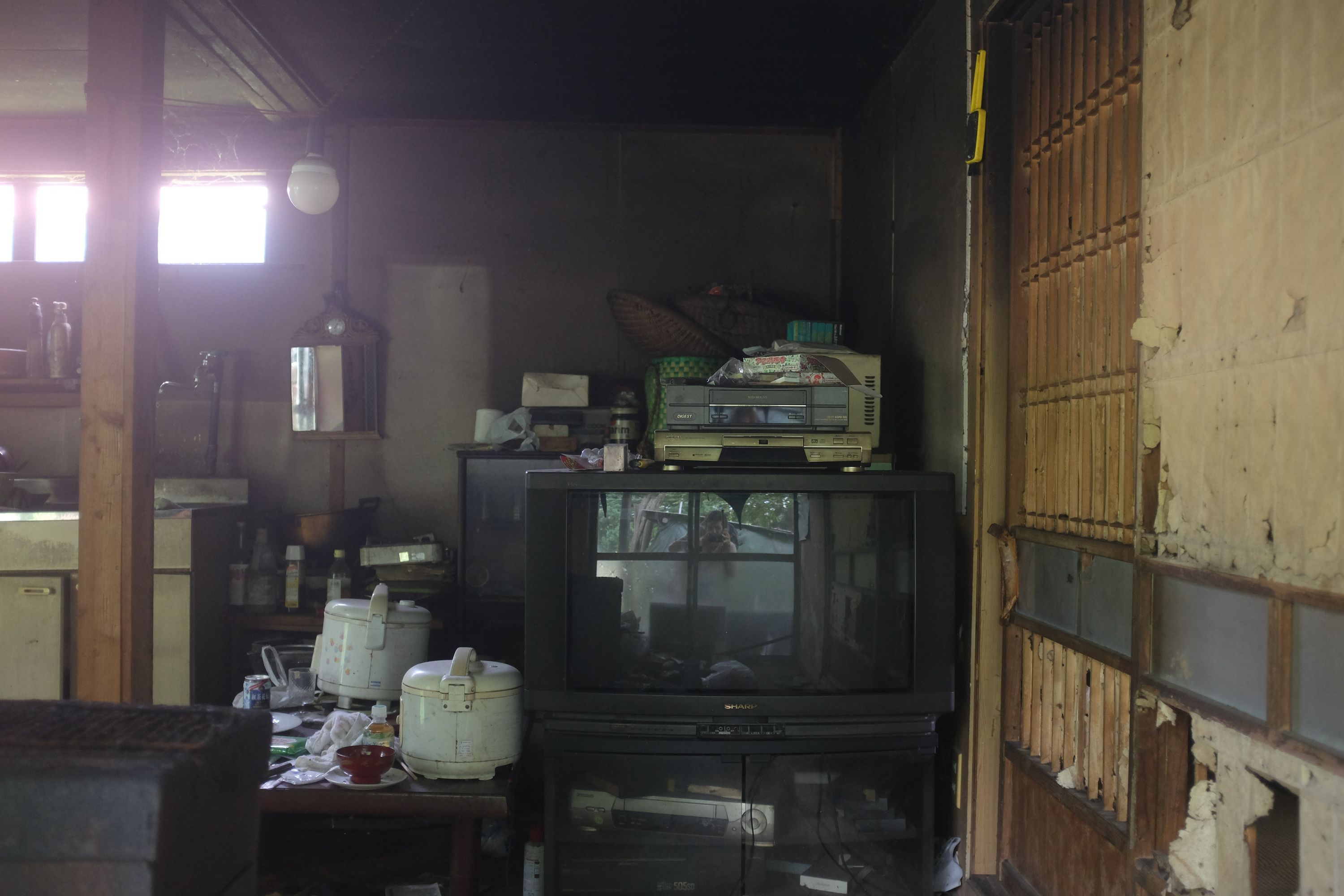 The kitchen of an abandoned house, showing appliances and kitchenware strewn about, with the author reflected in the screen of a television set.