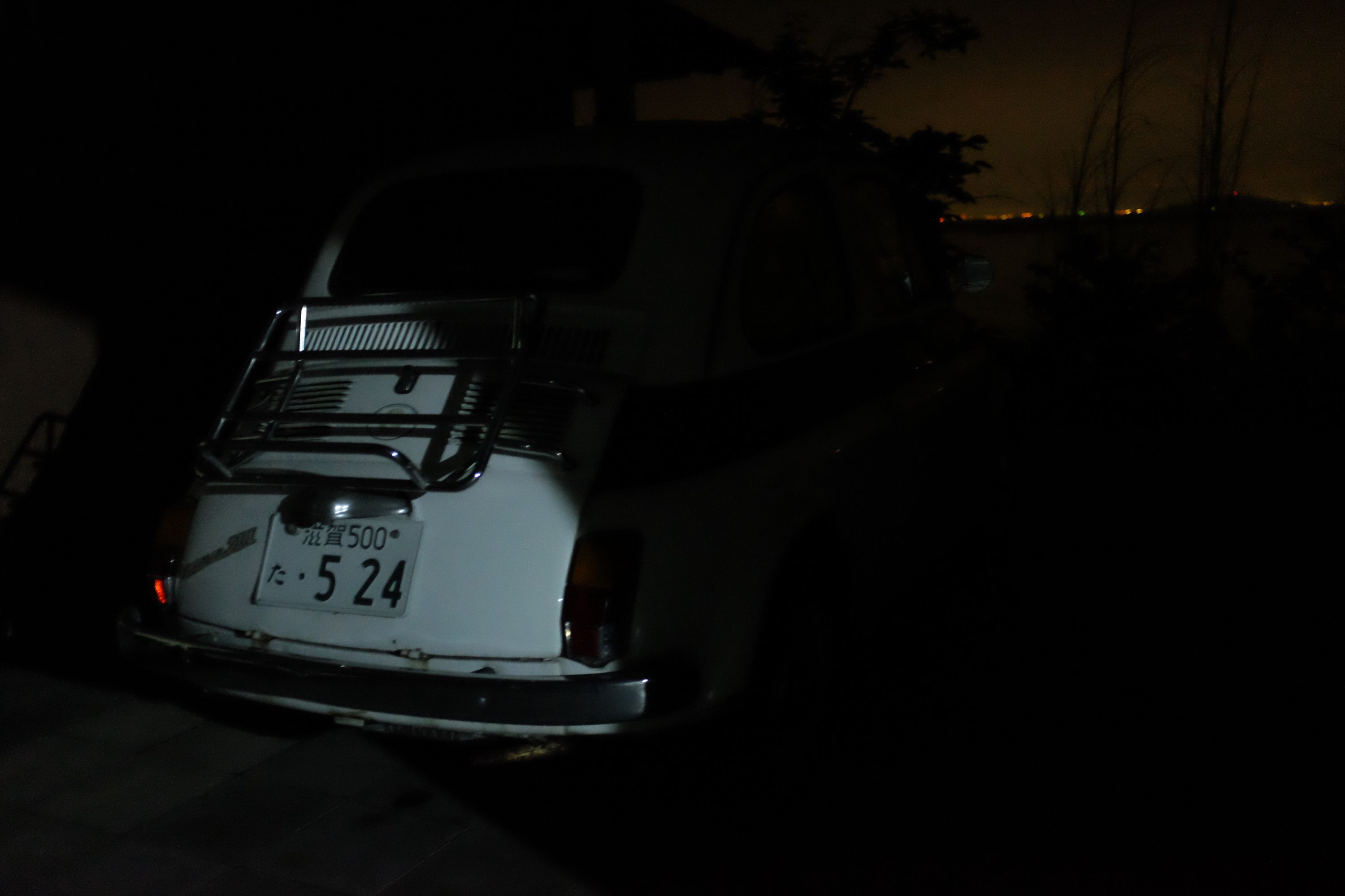 The rear end of a vintage white Fiat 500 is lit by a streetlamp, it is otherwise dark and there are distant lights on the horizon.