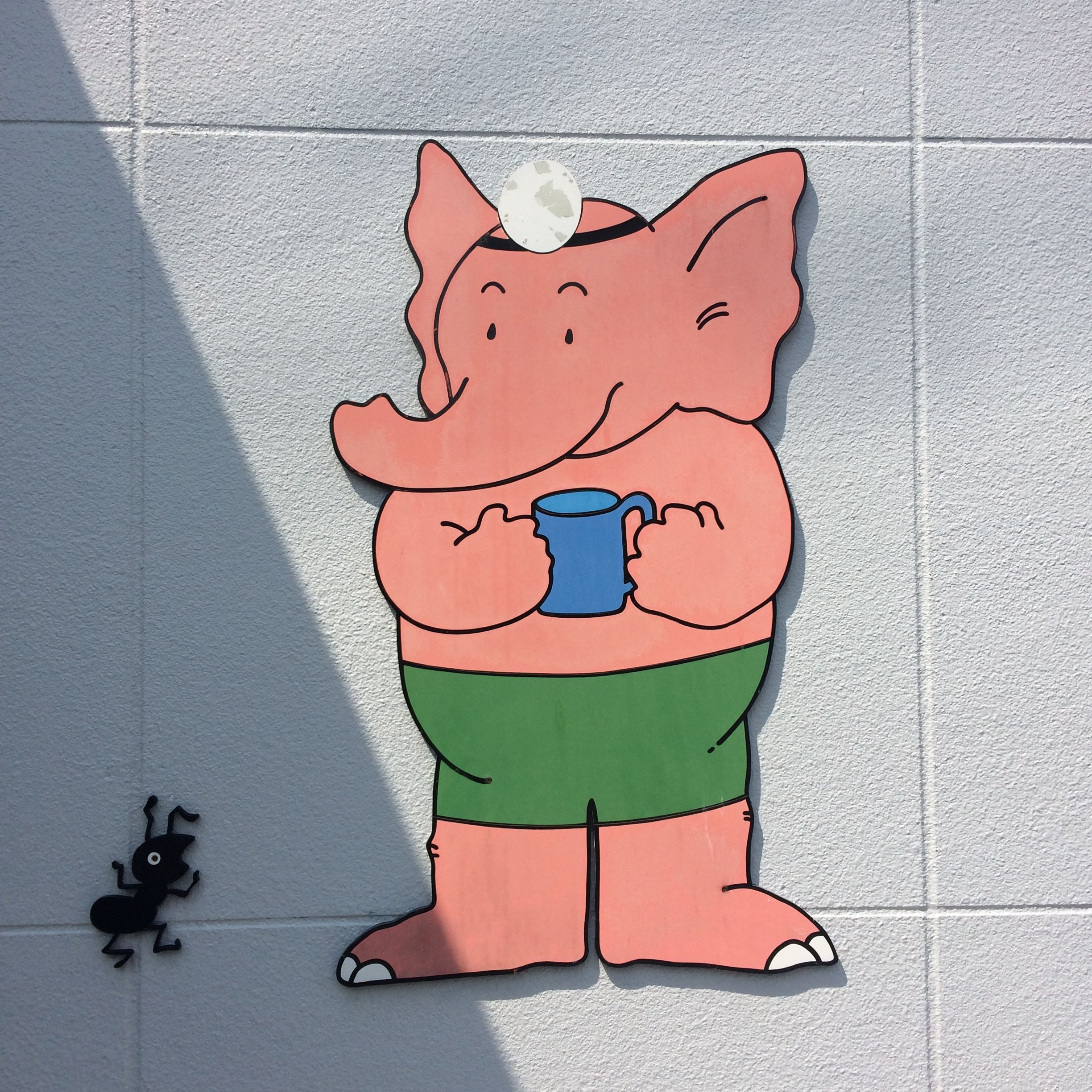 On the same wall as above, a cartoon elephant wearing green trunks and an ophthalmologist’s headlamp holds a blue mug and smiles at a small, worried ant.