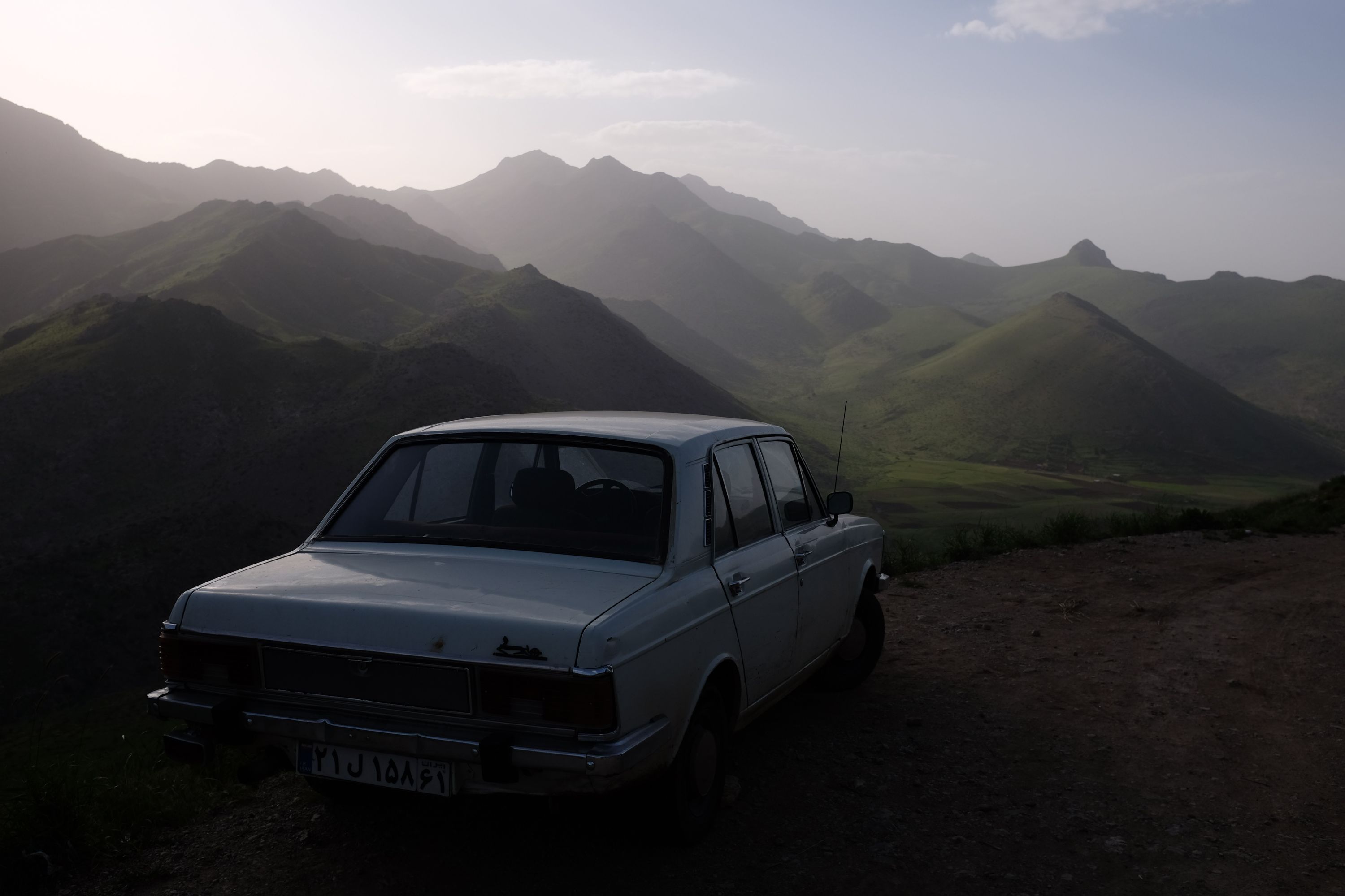 An old white sedan parked on a road with a view of rolling green hills.