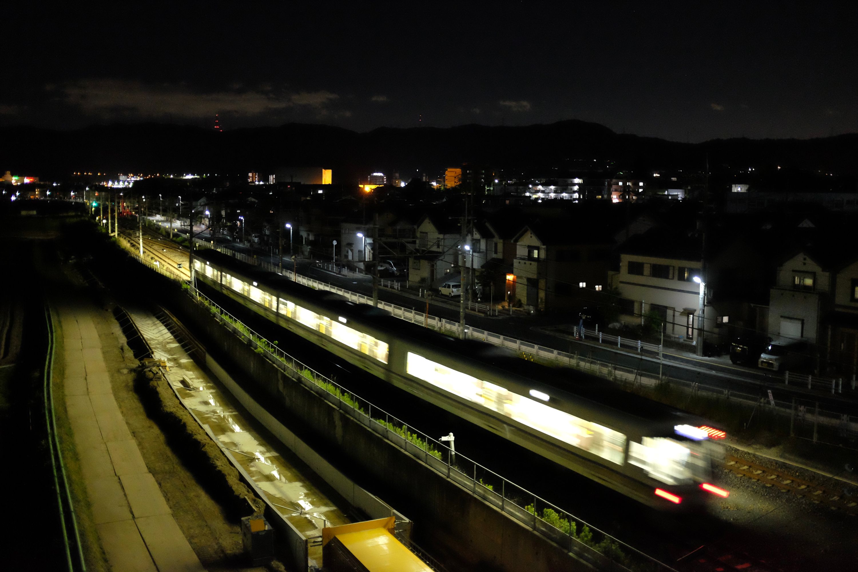 A train pulls into a station at night, against the lights of a city and low hills on the horizon.