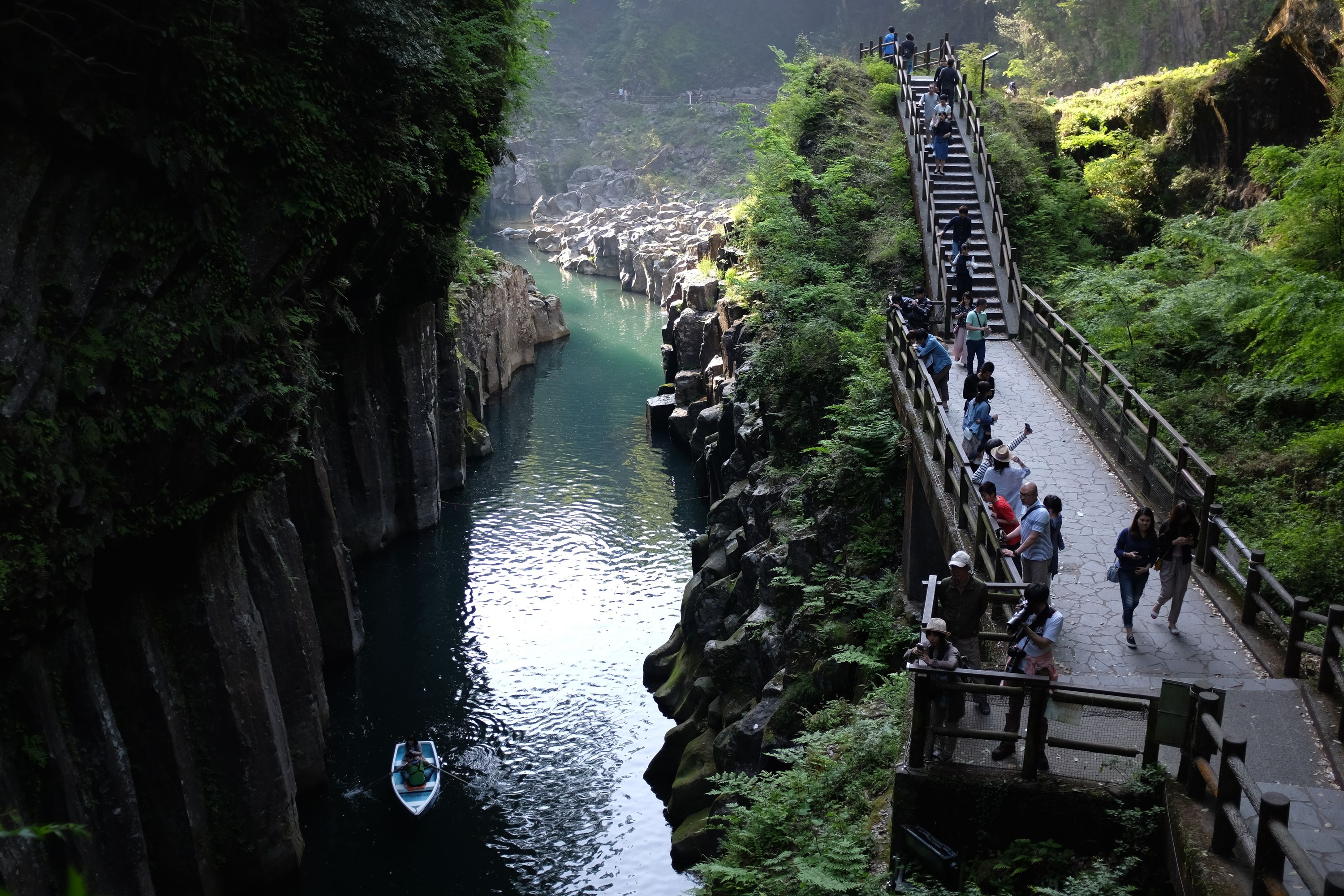 A lot of people on a walkway by a stream flowing through a gorge whose walls are black columns of basalt.